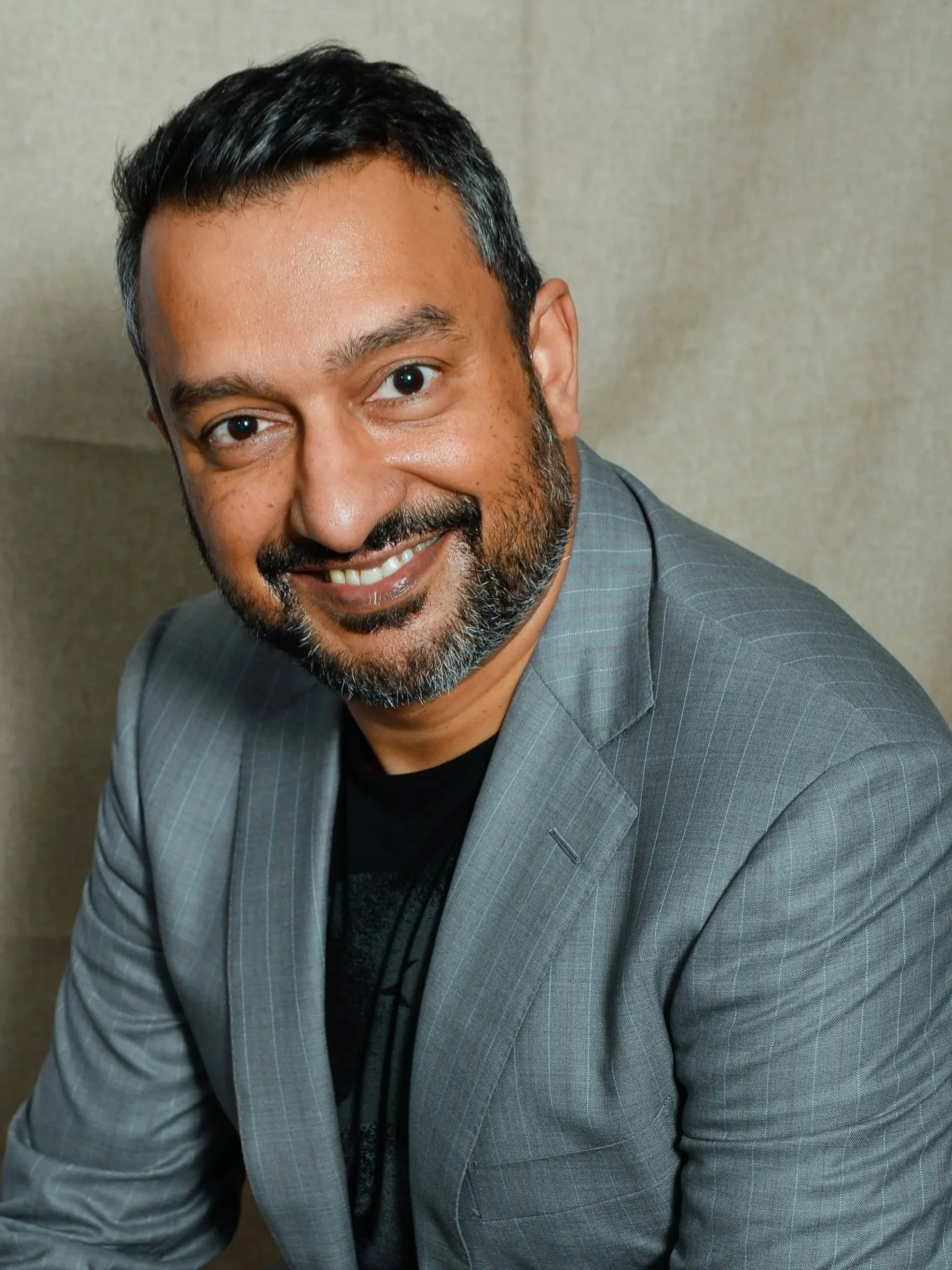 Close-up of a man with short dark hair, a beard, wearing a gray plaid suit, smiling in front of a neutral background.