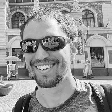 A man with curly hair and a beard wearing sunglasses and a t-shirt, smiling outdoors in front of a historic building with arched windows and decorative brickwork.