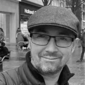 Man wearing glasses and a flat cap, smiling, in an urban setting.