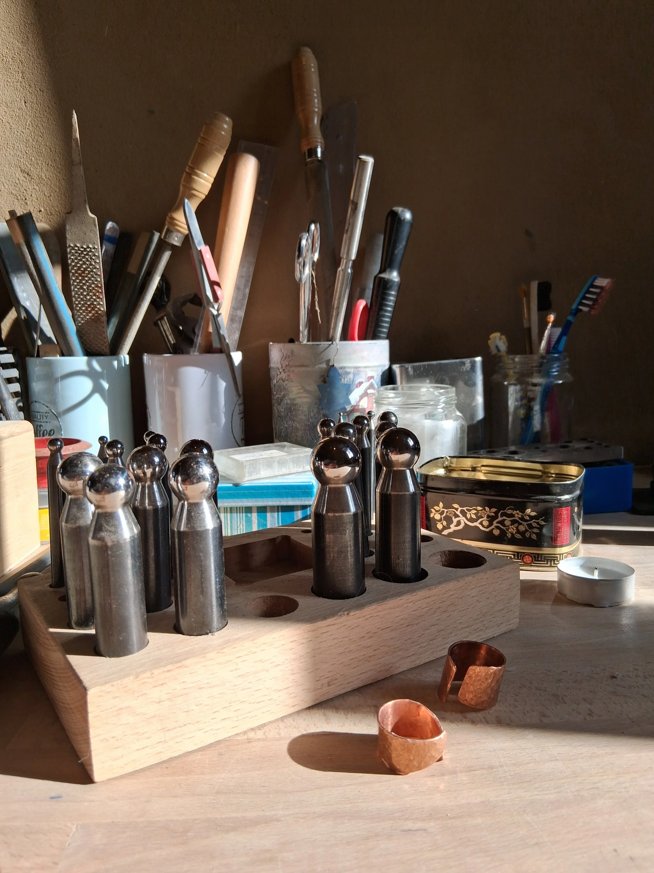 Ein Werktisch, vollgestellt mit verschiedenen Werkzeugen zur Schmuckherstellung, teils liegend, teils in Gefäßen verstaut, auf einer Arbeitsplatte und vor einer braunen Lehmwand.