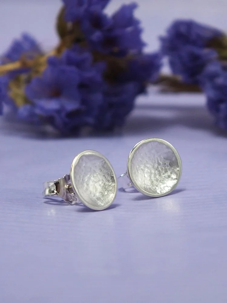 Silberne Ohrringe mit rundem, leicht strukturiertem Schalendesign, im Hintergrund getrocknete Blüten auf violettem Hintergrund.