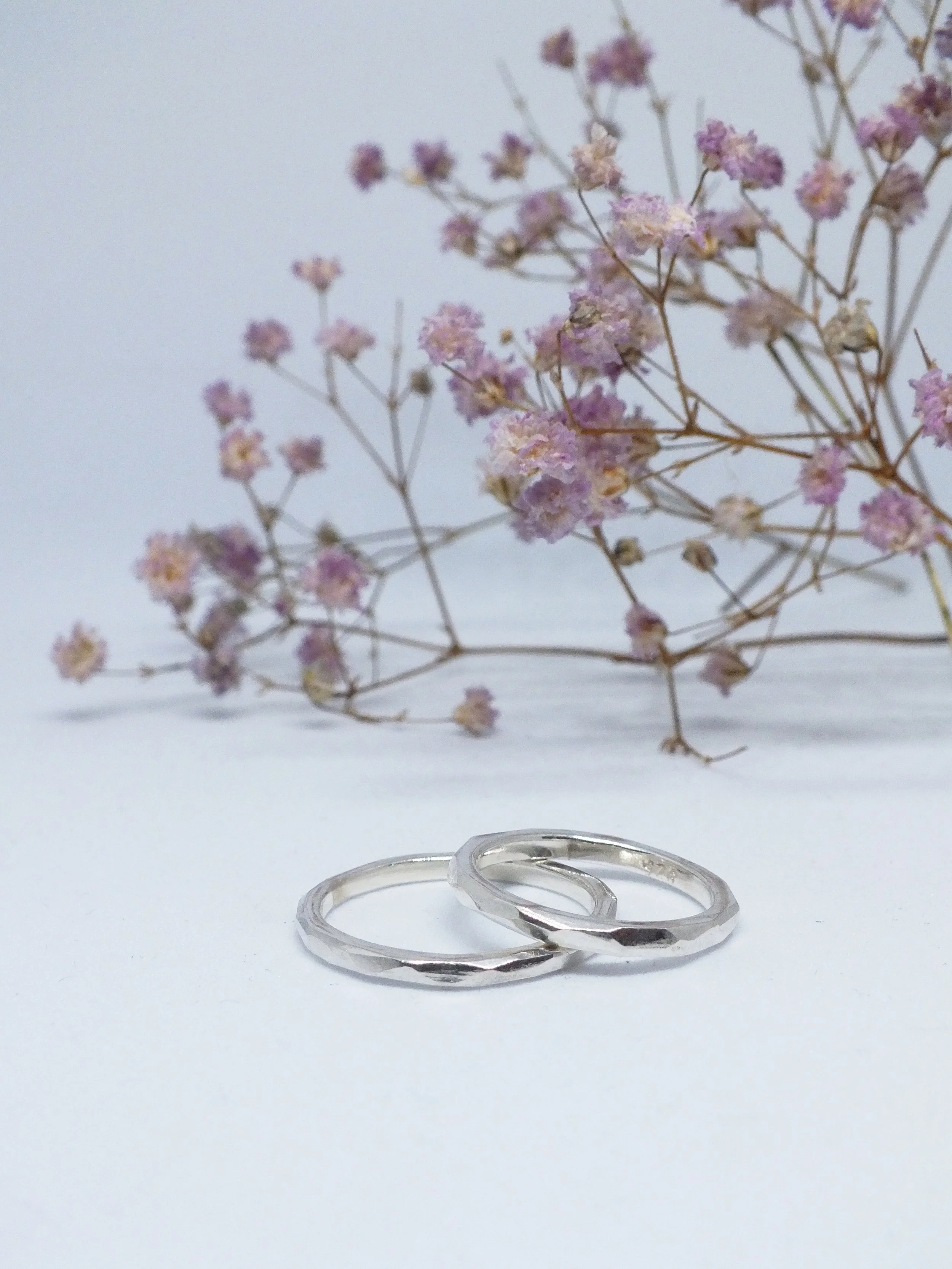 Zwei geschichtete silberne Ringe mit angefeilten Facetten vor einem Hintergrund mit lila Blumen, auf weißem Untergrund.
