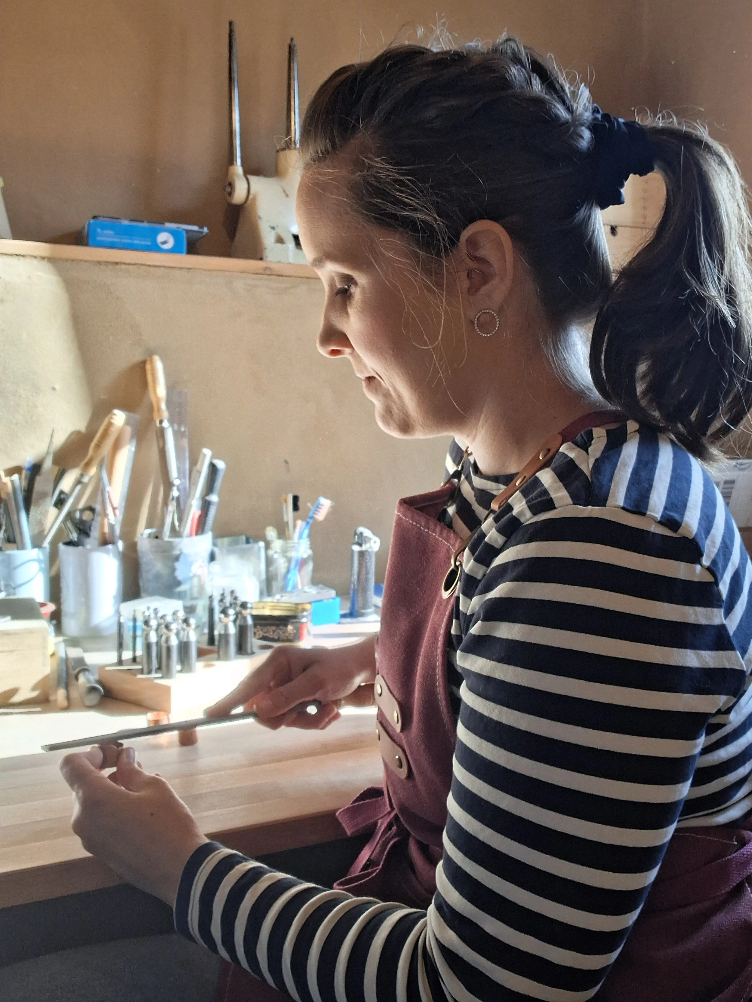 Frau arbeitet in einer Werkstatt mit Feile und Werkzeugen an einem Werktisch, trägt eine dunkelrote Schürze und hat dunkles, geflochtenes Haar, das zu einem Pferdeschwanz gebunden ist.