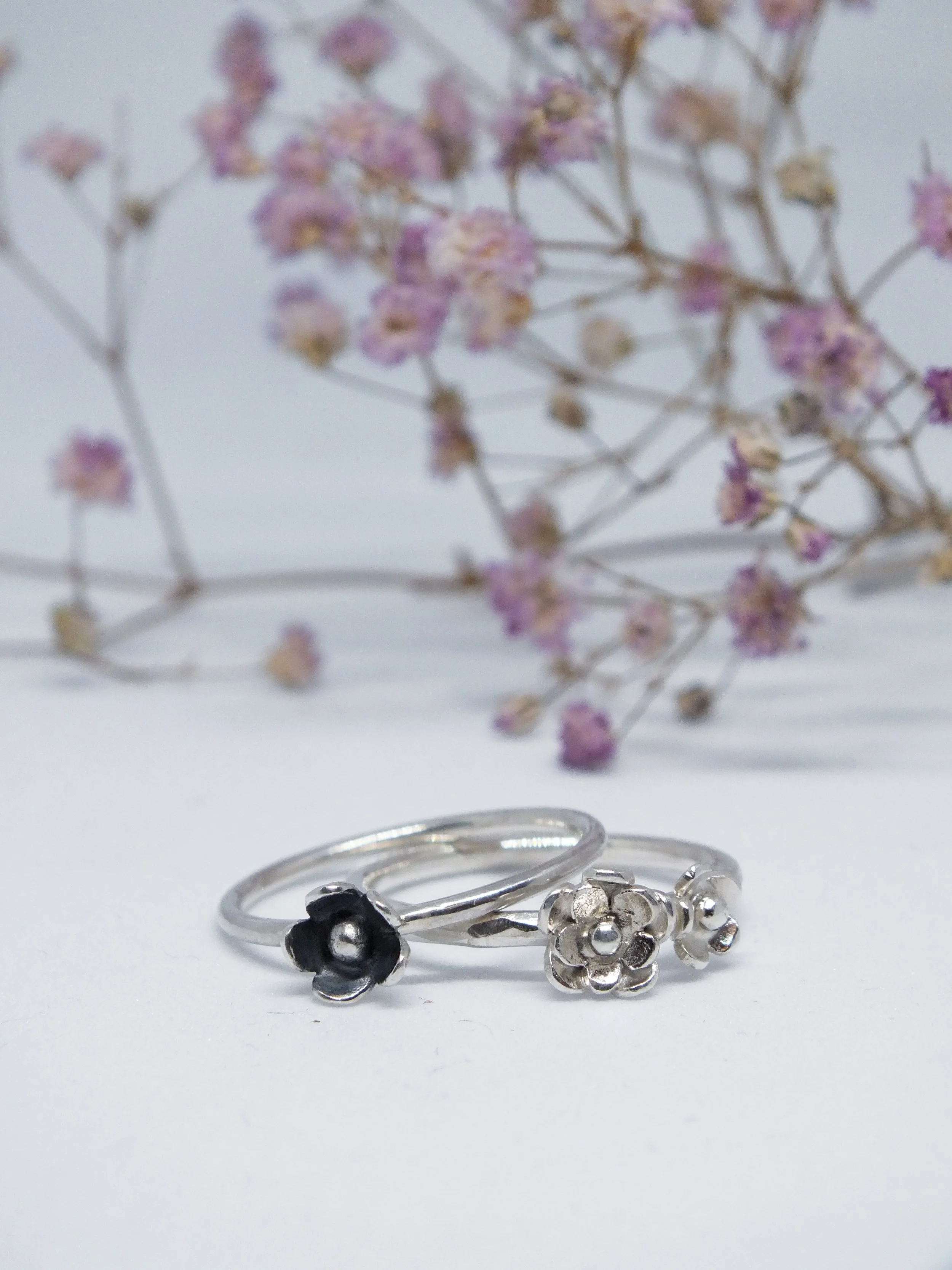 Zwei Silberringen mit Blumendesign, vor einem Zweig mit kleinen, lila Blüten auf weißem Hintergrund.