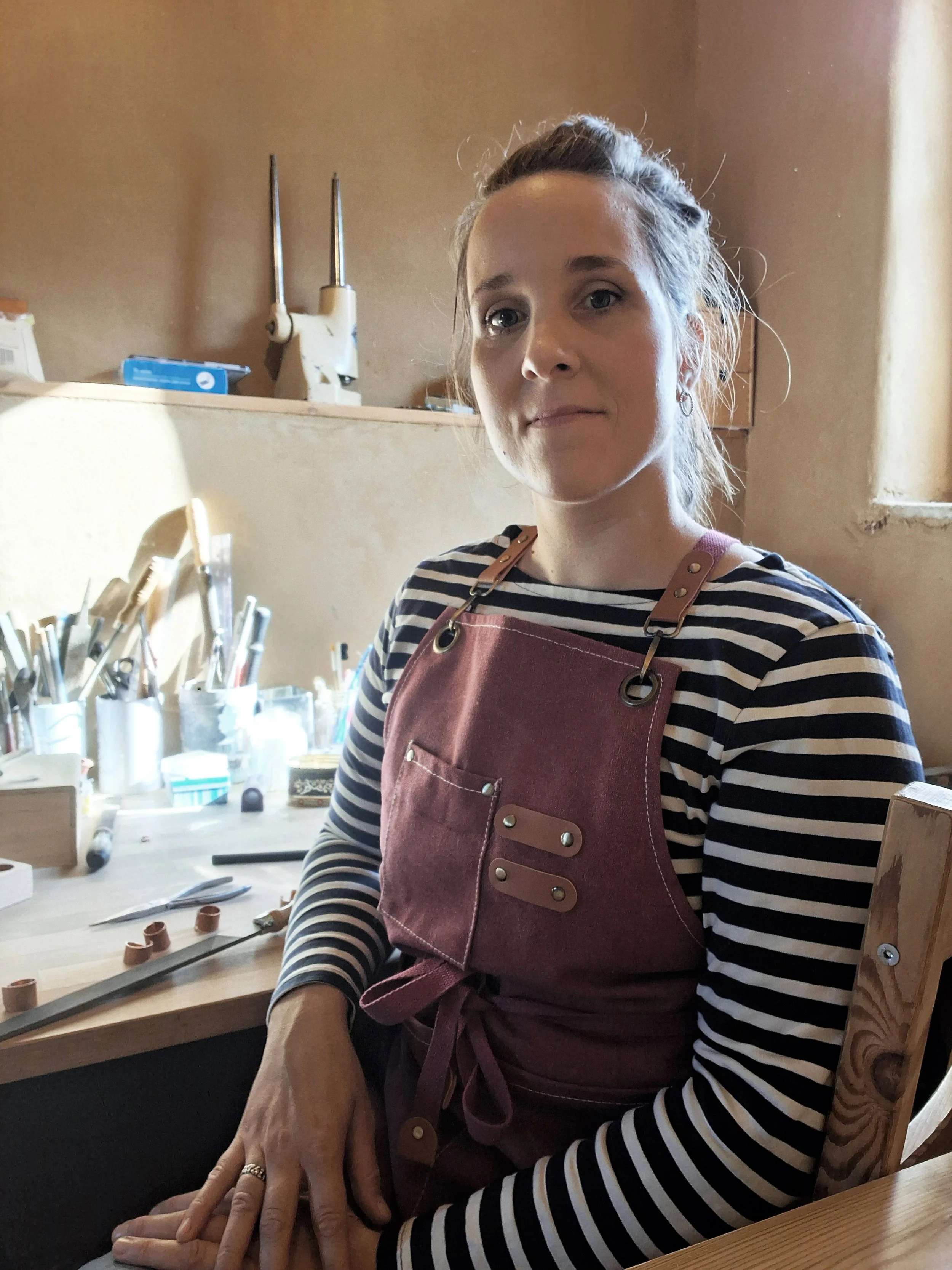 Junge Frau in einer Werkstatt trägt einer dunkelroten Schürze mit Ledergriffen, sitzt an einem Tisch mit Werkzeug und lehnte an einem Holzstuhl.