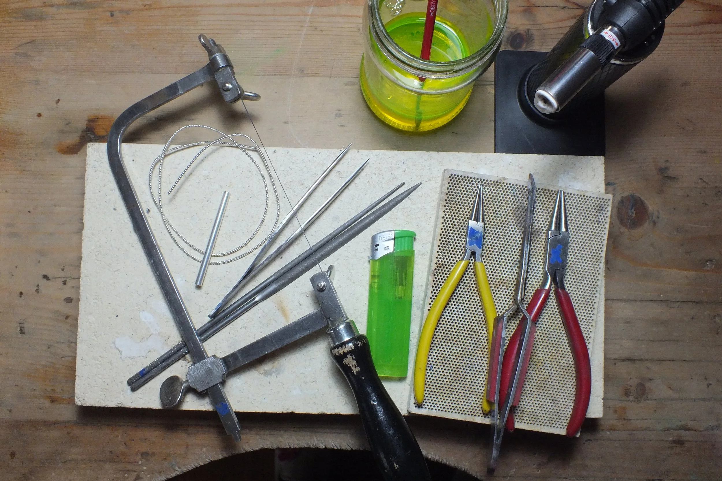 Blick auf einen Werkzeittisch mit verschiedenen Werkzeugen, Draht, Zange, Messer, Löffel, viel Zubehör, ein Feuerzeug, Kerzendocht, eine kleine Aluhülle, und ein Glas mit grünem Getränk und Strohhalm.