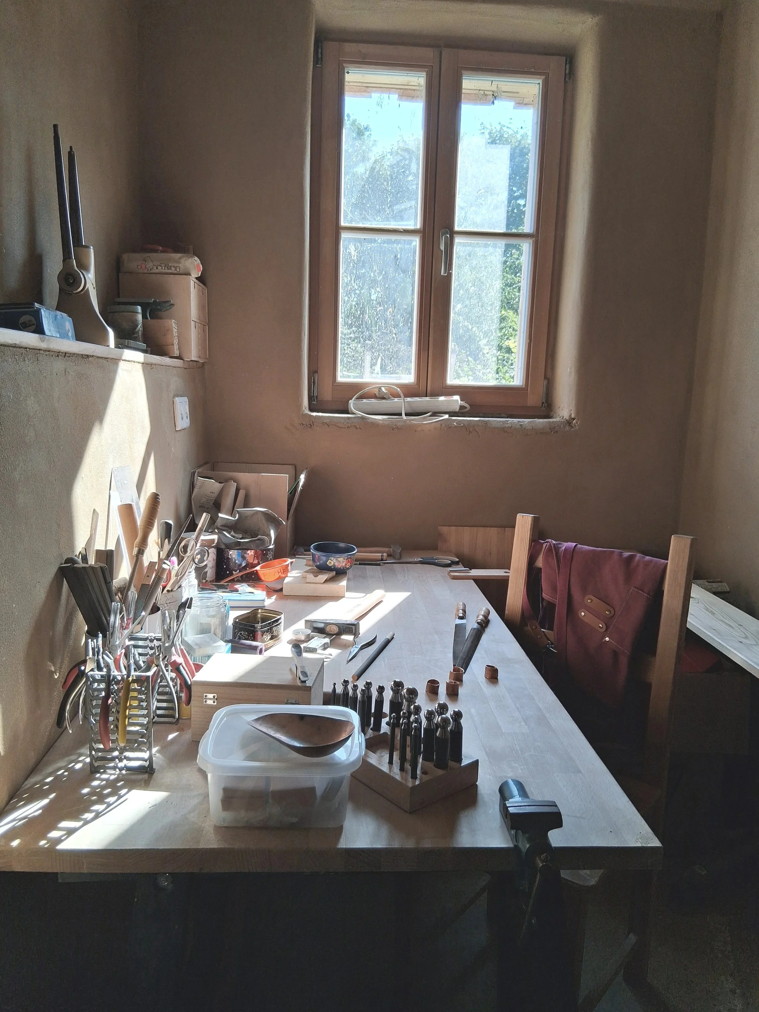 Ein Arbeitsplatte mit Werkzeugen und Materialien, eine Fenster mit Blick nach draußen, und ein Stuhl mit einem braunen Mantel darauf in einem Raum bei Tageslicht.