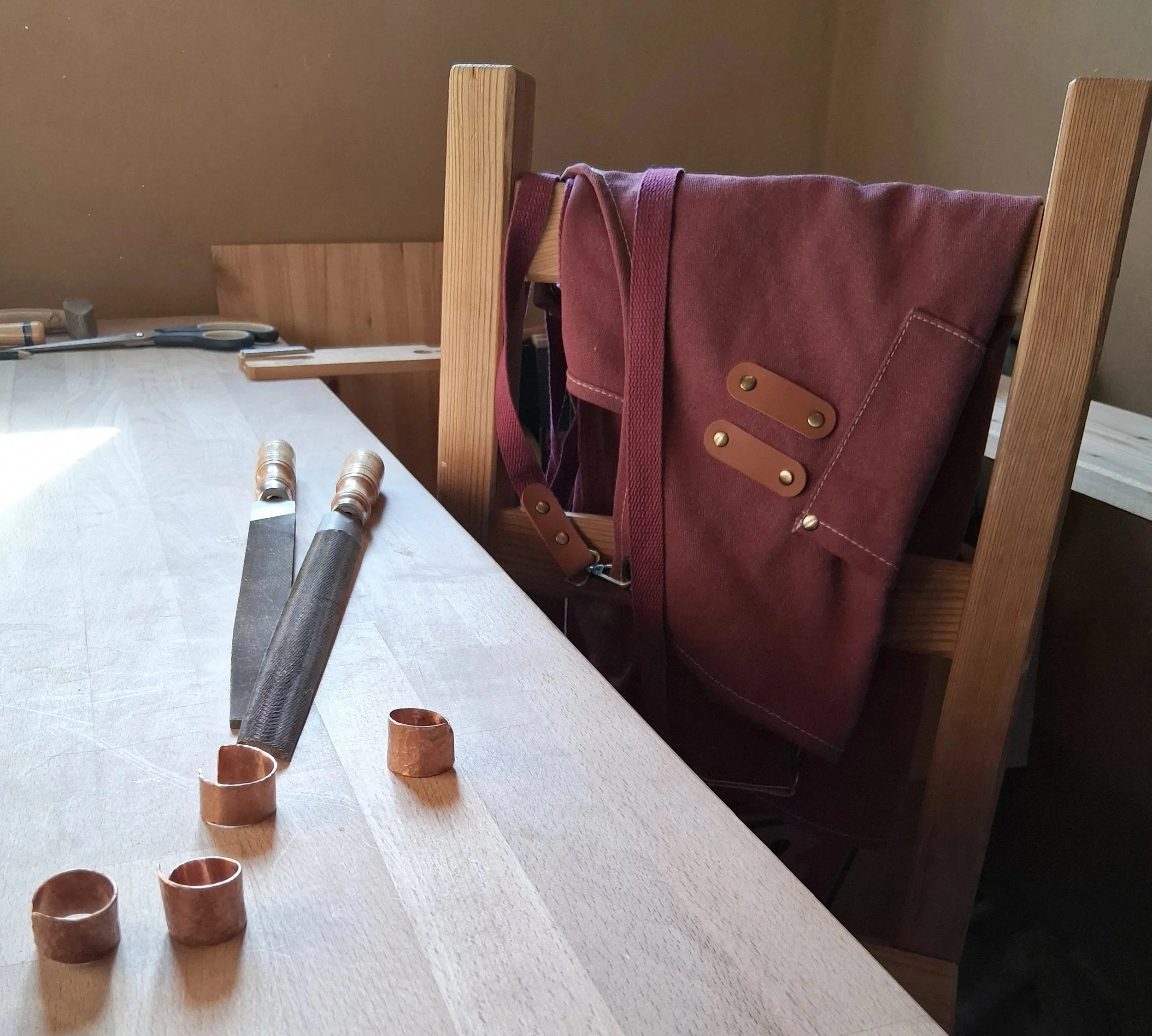 Feilen und Ringkörper aus Kupfer auf einem Werktisch mit einer roten Schürze auf einem Stuhl in einer Werkstattumgebung.