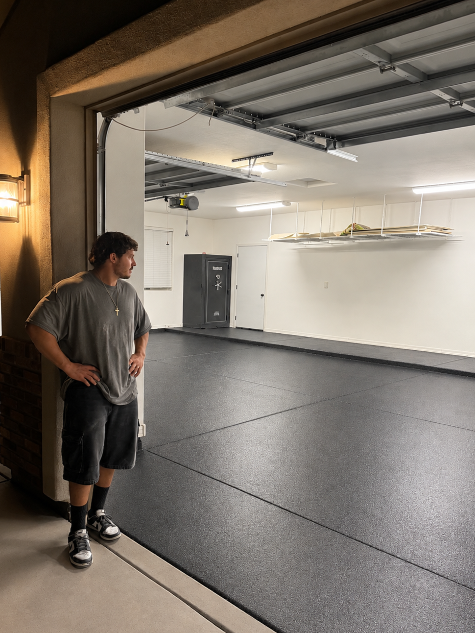 A young man standing near the egress doorway of a garage, looking inside at an empty, clean, lighted garage with black flooring and some stored items on a shelf.