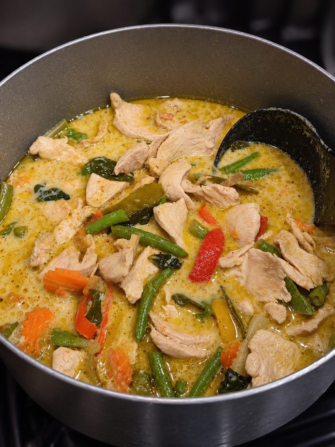 Chicken and vegetable curry cooking in a pot with a ladle, including chicken pieces, red and green bell peppers, carrots, and green beans in a yellow curry sauce.