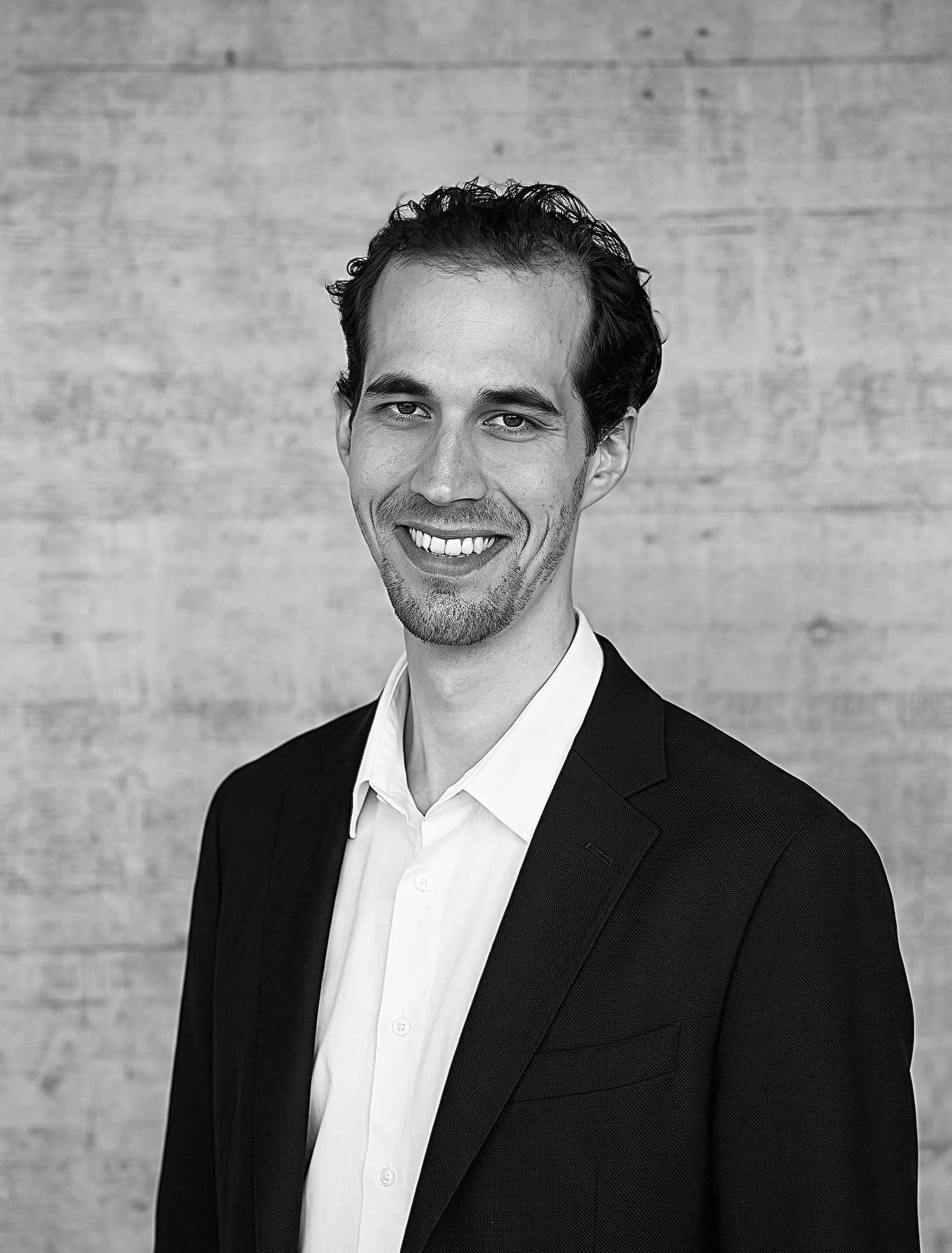 Schwarz-Weiss-Porträt von Urs Büchi, Projektleiter Architektur und Baurecht der Innovaplus integrale Planung GmbH, im Anzug vor einer Sichtbetonwand.