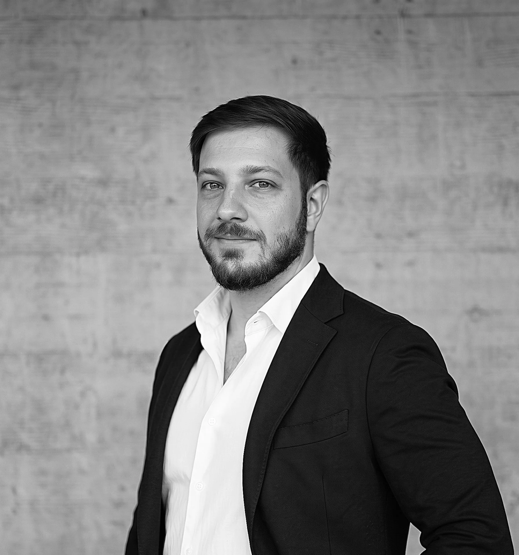 Schwarz-Weiss-Porträt von Luca Palmisano, Gesamtprojektleiter und Geschäftsleitung der Innovaplus integrale Planung GmbH, im Anzug vor einer Sichtbetonwand.