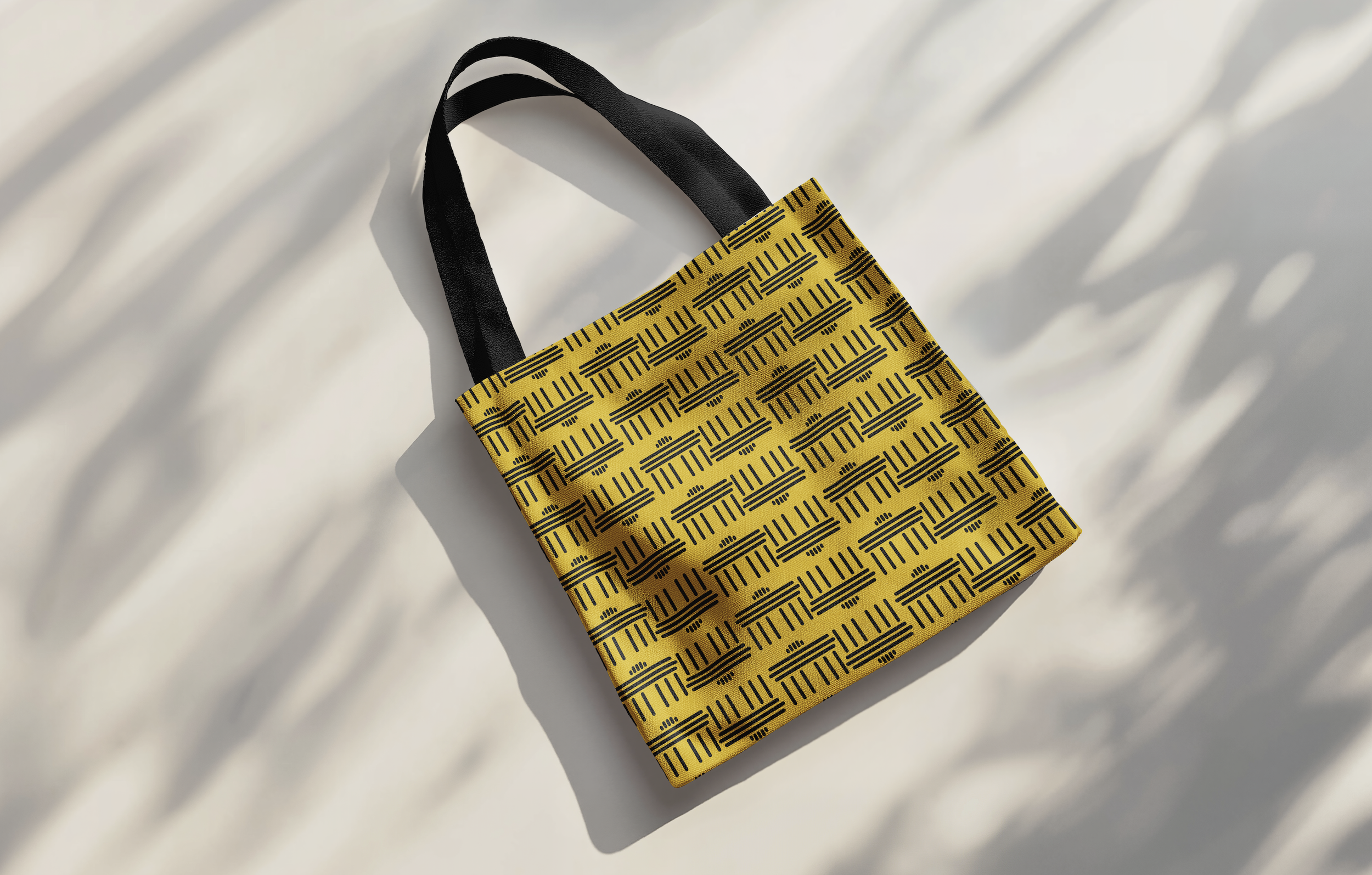 Gelbe Stofftasche mit schwarzen geometrischen Linienmustern und schwarzen Tragegriffen, liegt auf weißem Hintergrund mit Schatten.