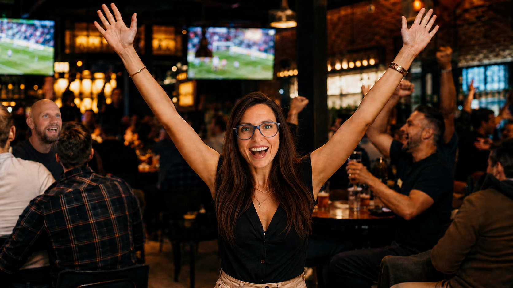 A woman with glasses celebrating with her arms raised in a crowded sports bar, watching a game on multiple large screens.