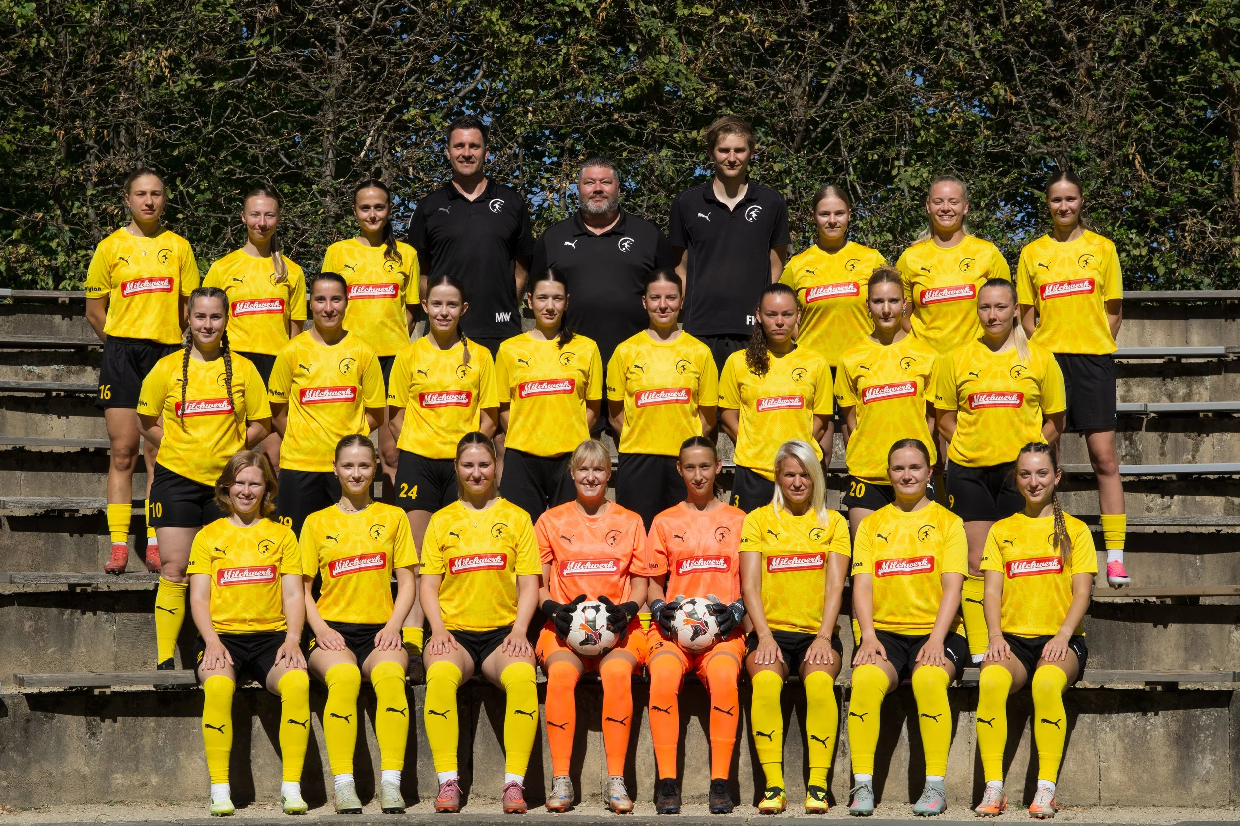 Ein Fußballteam bestehend aus Mädchen in gelben und orangefarbenen Trikots sowie drei Trainer oder Betreuer steht auf Stufen vor einer Baumwand. Das Foto ist bei schönem Wetter aufgenommen.