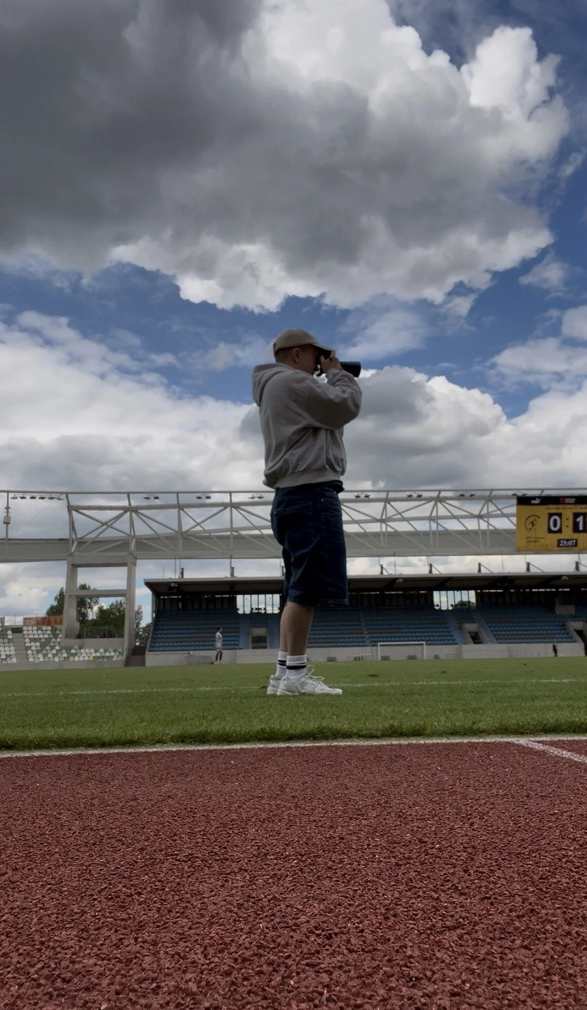 Ein junger Mann steht auf einer Leichtathletikbahn auf einem Sportplatz und schaut durch ein Fernglas in die Ferne, während Wolken am Himmel sind.