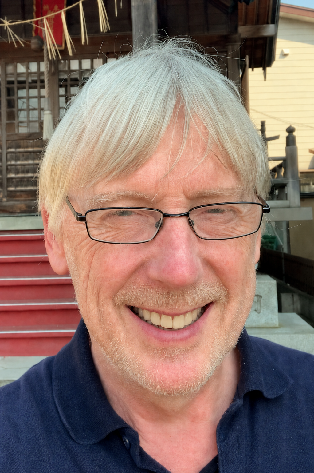 A smiling man with glasses and white hair standing outside in front of a shrine with shimenawa rope and shide paper streamers.