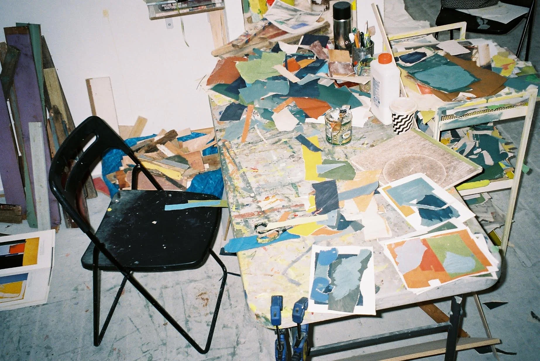 A cluttered art studio workspace with paint color samples, scattered papers, art supplies, and a black chair.