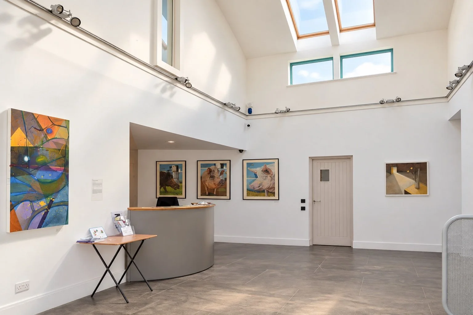 Bright Moray Art Centre atrium with skylights, colourful artwork, reception desk, framed paintings, tiled floor and an entrance doorway.