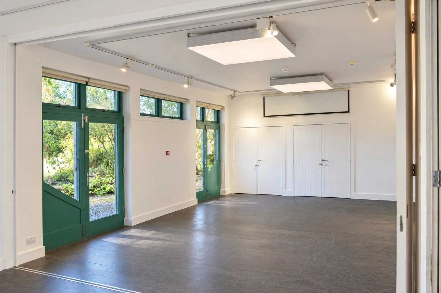 Garden-facing classroom with green doors, projector screen and open floor.