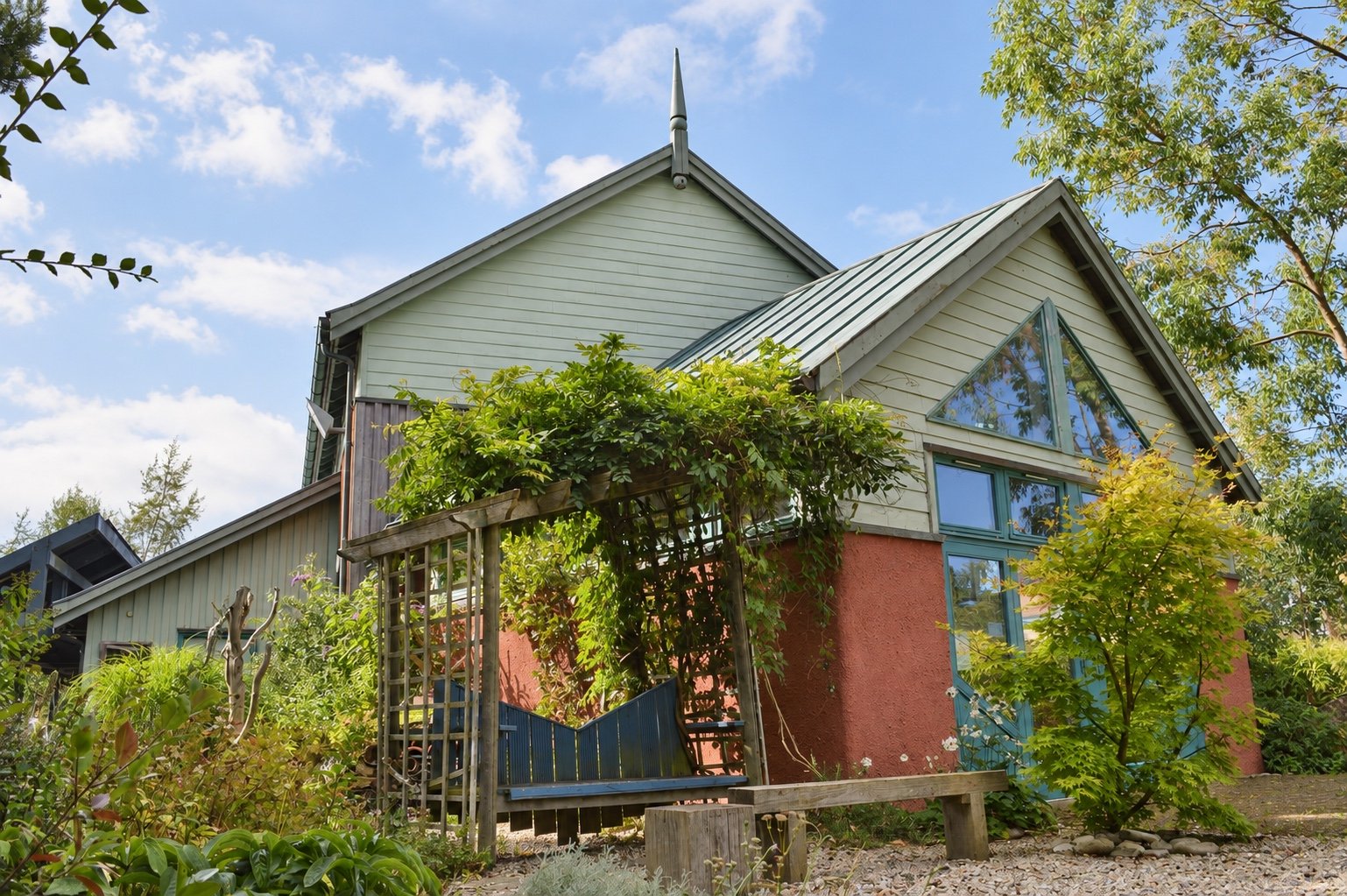 Exterior of Moray Art Centre Classroom C with pale timber gables, green-framed windows, a vine-covered trellis, blue gate, bench and planted garden.