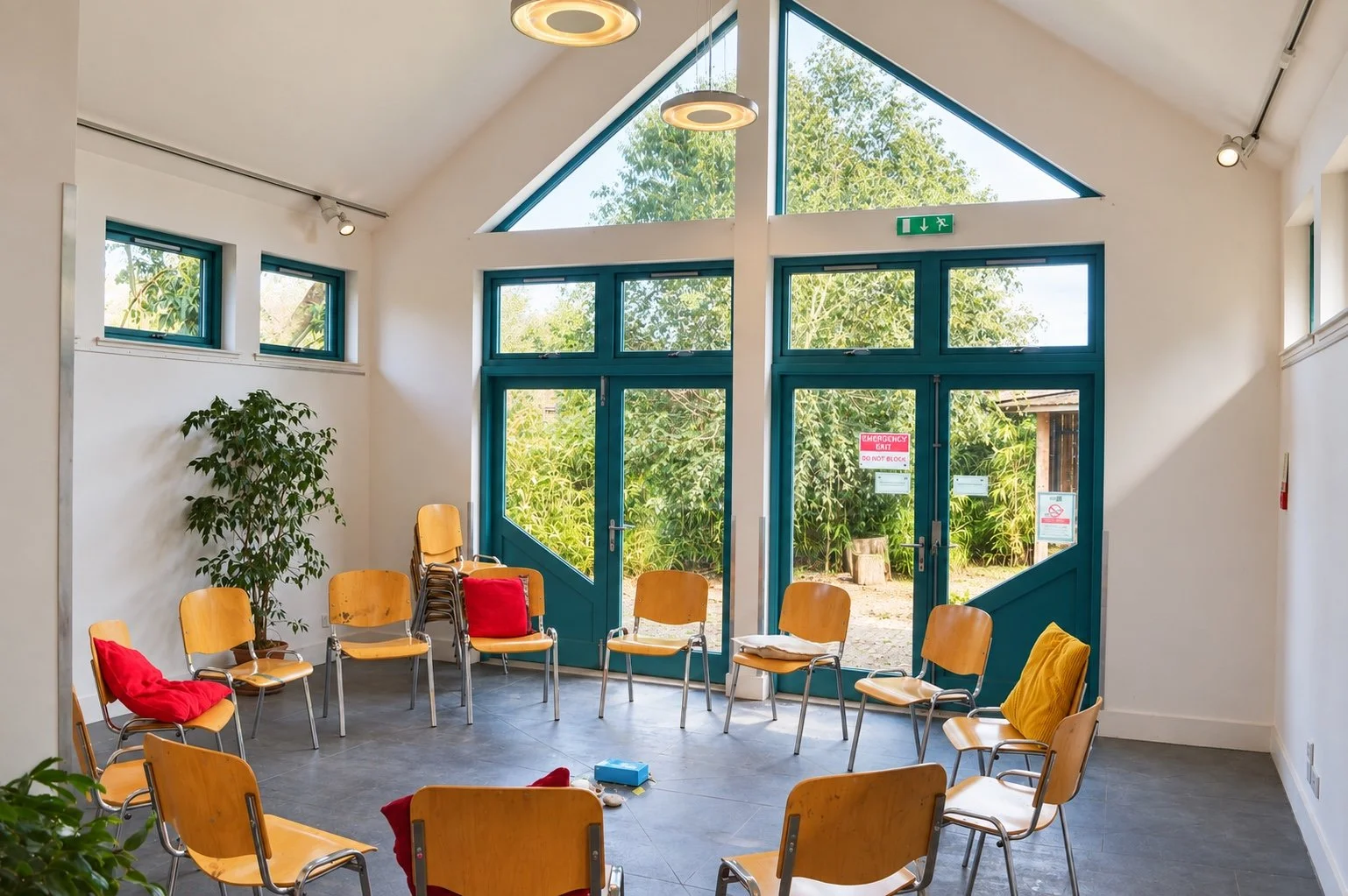 Bright Moray Art Centre gathering room with chairs arranged in a circle, teal-framed glass doors, triangular windows, and a leafy garden beyond.