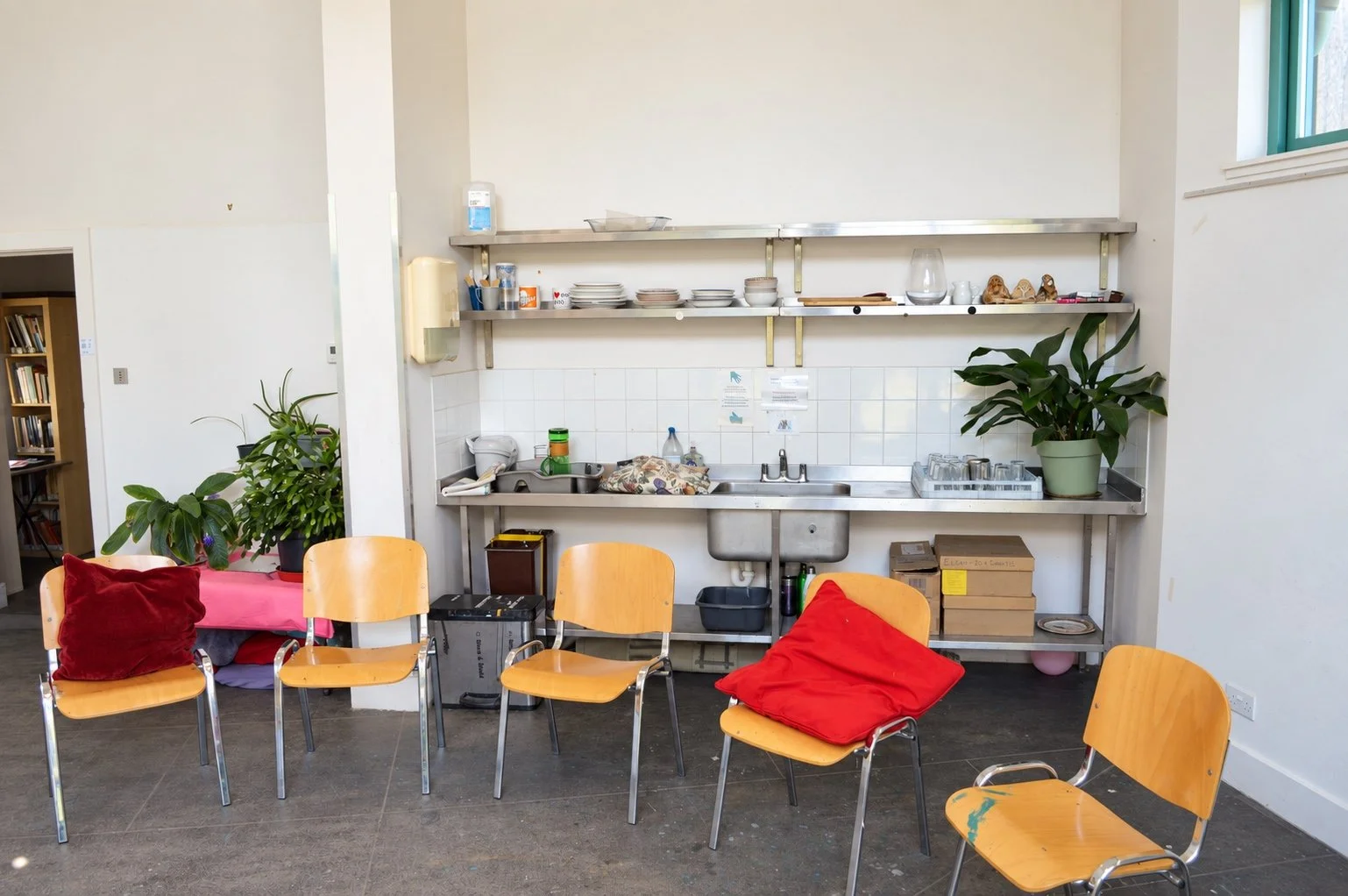Interior of Moray Art Centre Classroom C with chairs, plants, open shelving holding crockery, and a stainless-steel sink and work surface.