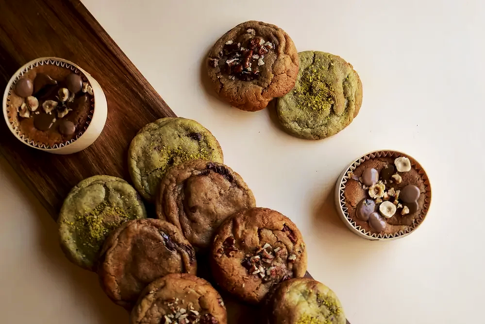 Cookies moelleux aux pistaches, chocolat et noisettes sur une table en bois et une surface blanche.