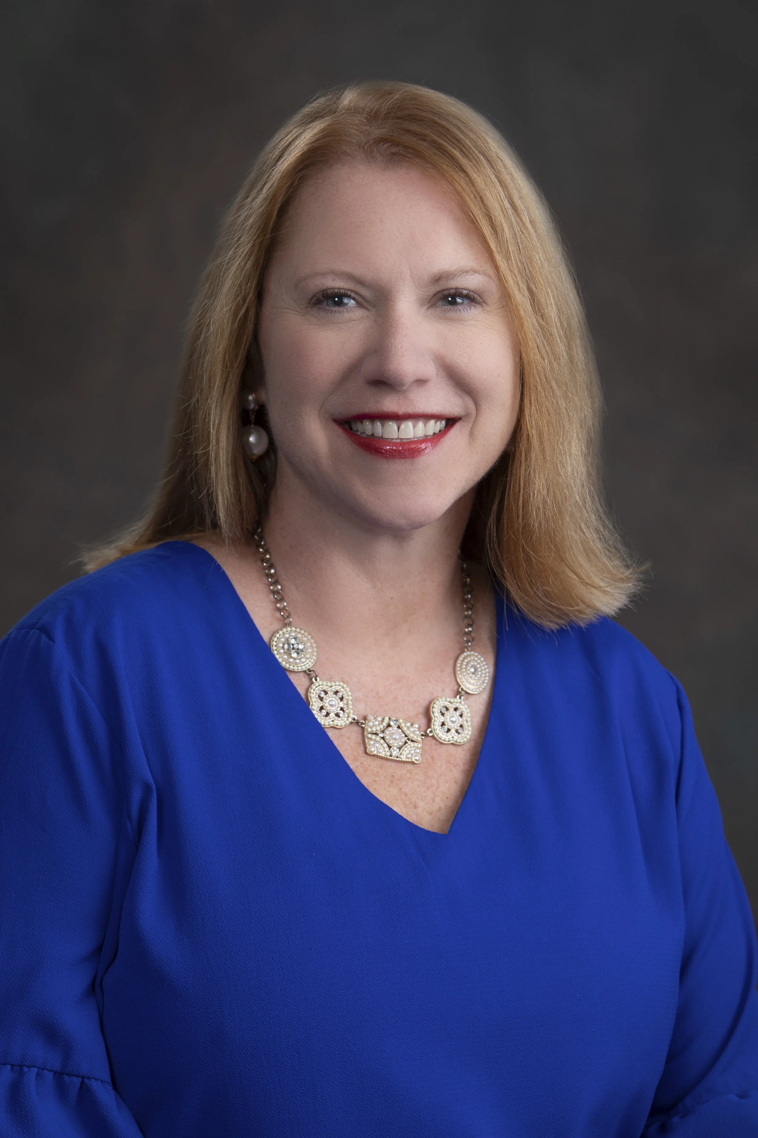 Woman with red hair wearing a blue top, pearl earrings, and a pearl necklace, smiling, against a dark background.