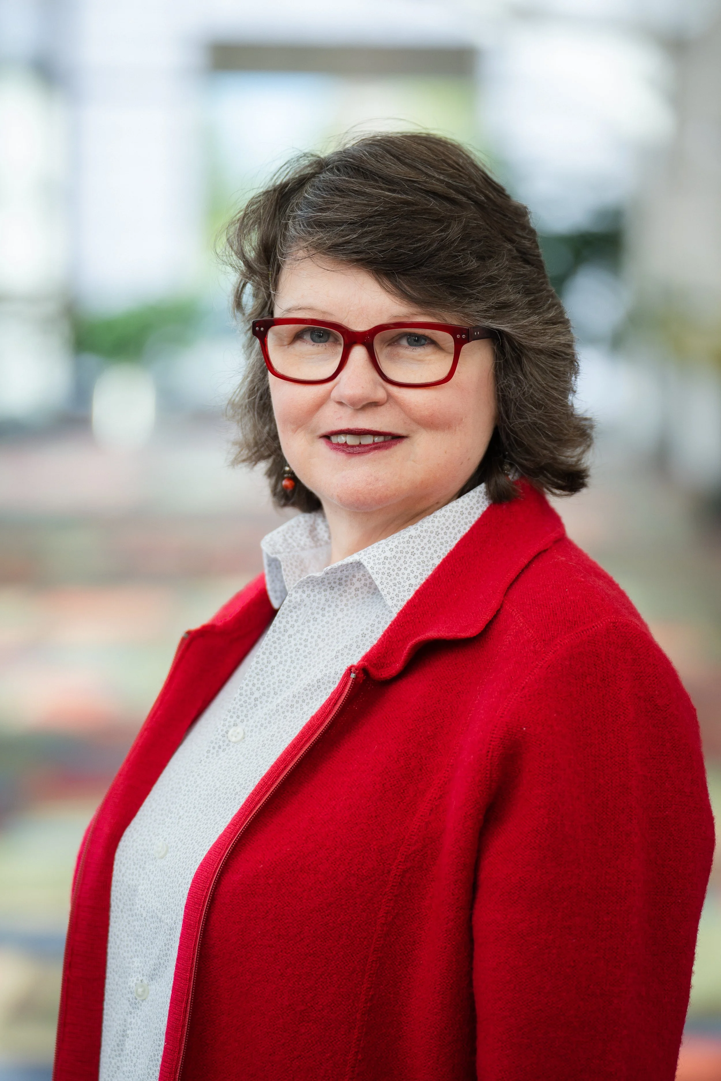 A woman with short, curly gray hair wearing red glasses, a white collared shirt, and a red sweater, smiling confidently outdoors.