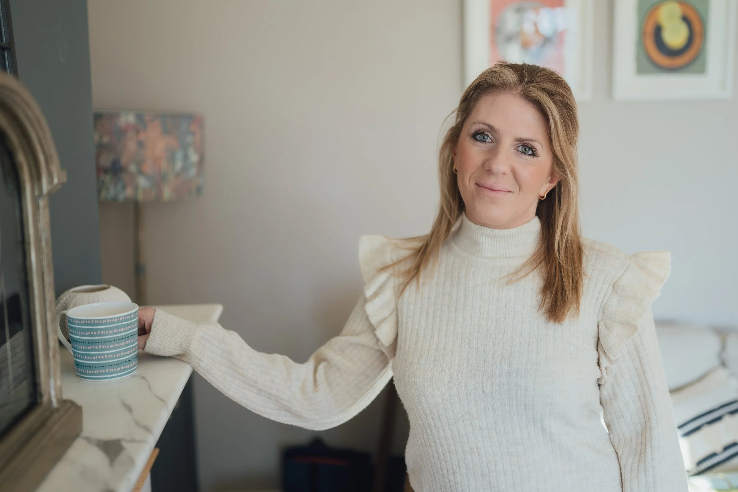 A woman with red hair and blue eyes wearing a cream-colored sweater, holding a blue and white striped coffee mug, standing in a kitchen with a marble countertop and colorful artwork on the wall in the background.