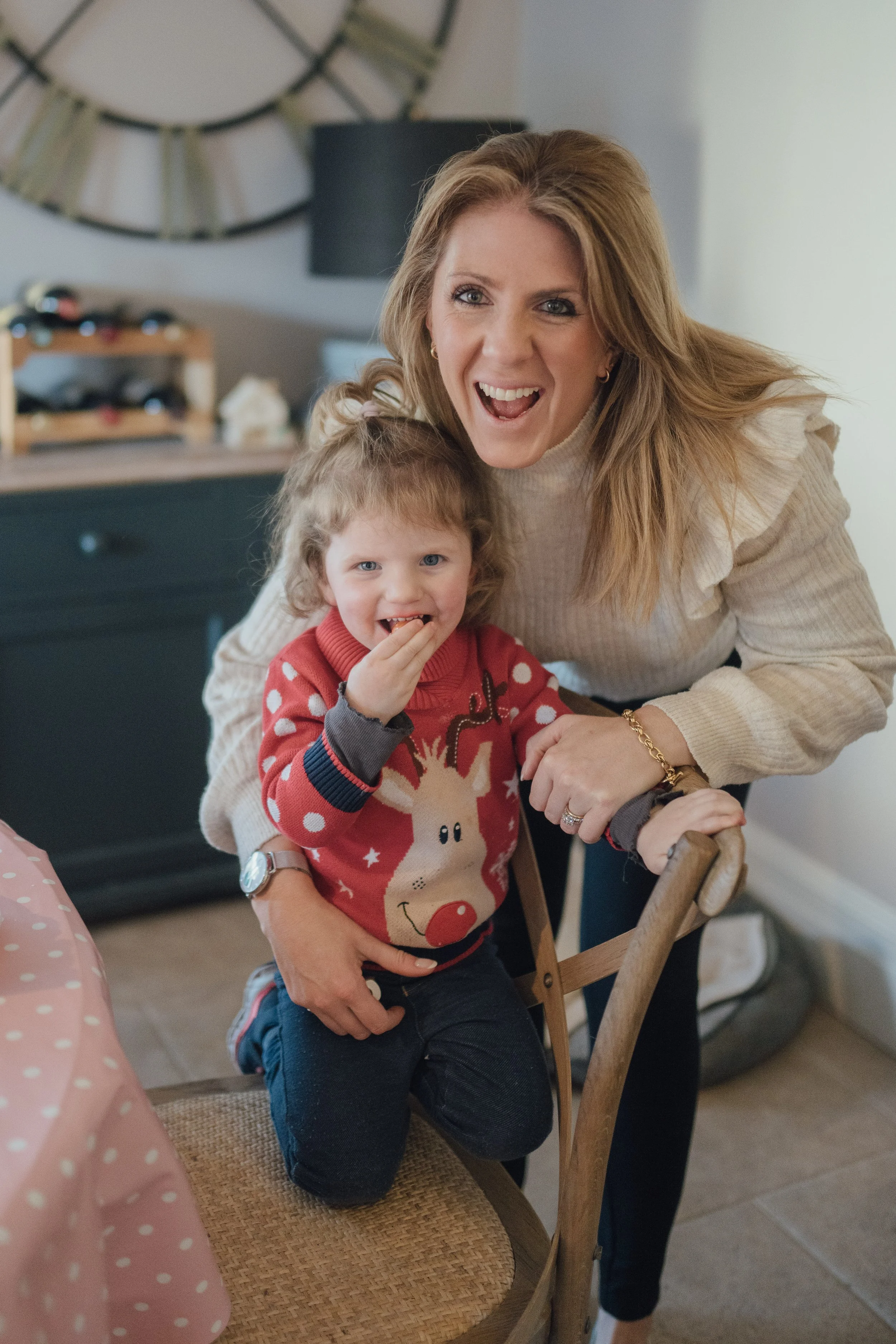 A woman with light brown hair and blue eyes smiling while holding a young girl with curly light brown hair and blue eyes, sitting on a wooden chair in a home interior.
