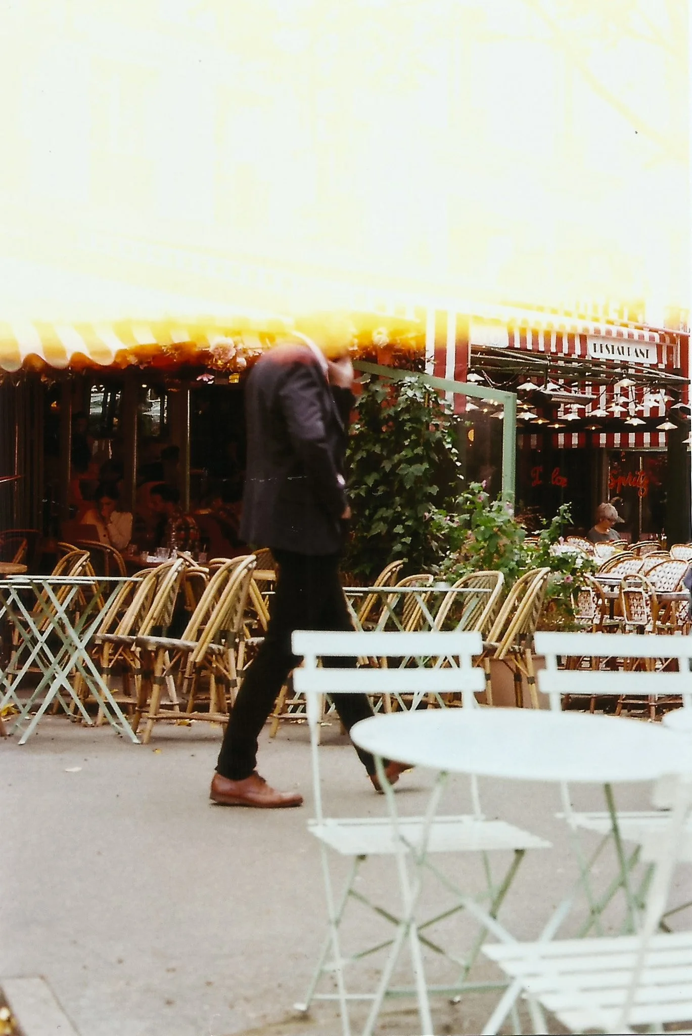Un homme habillé en costume marche dans une rue extérieure d'un café ou restaurant avec des chaises vides en avant-plan, des tables et des parasols colorés en arrière-plan.