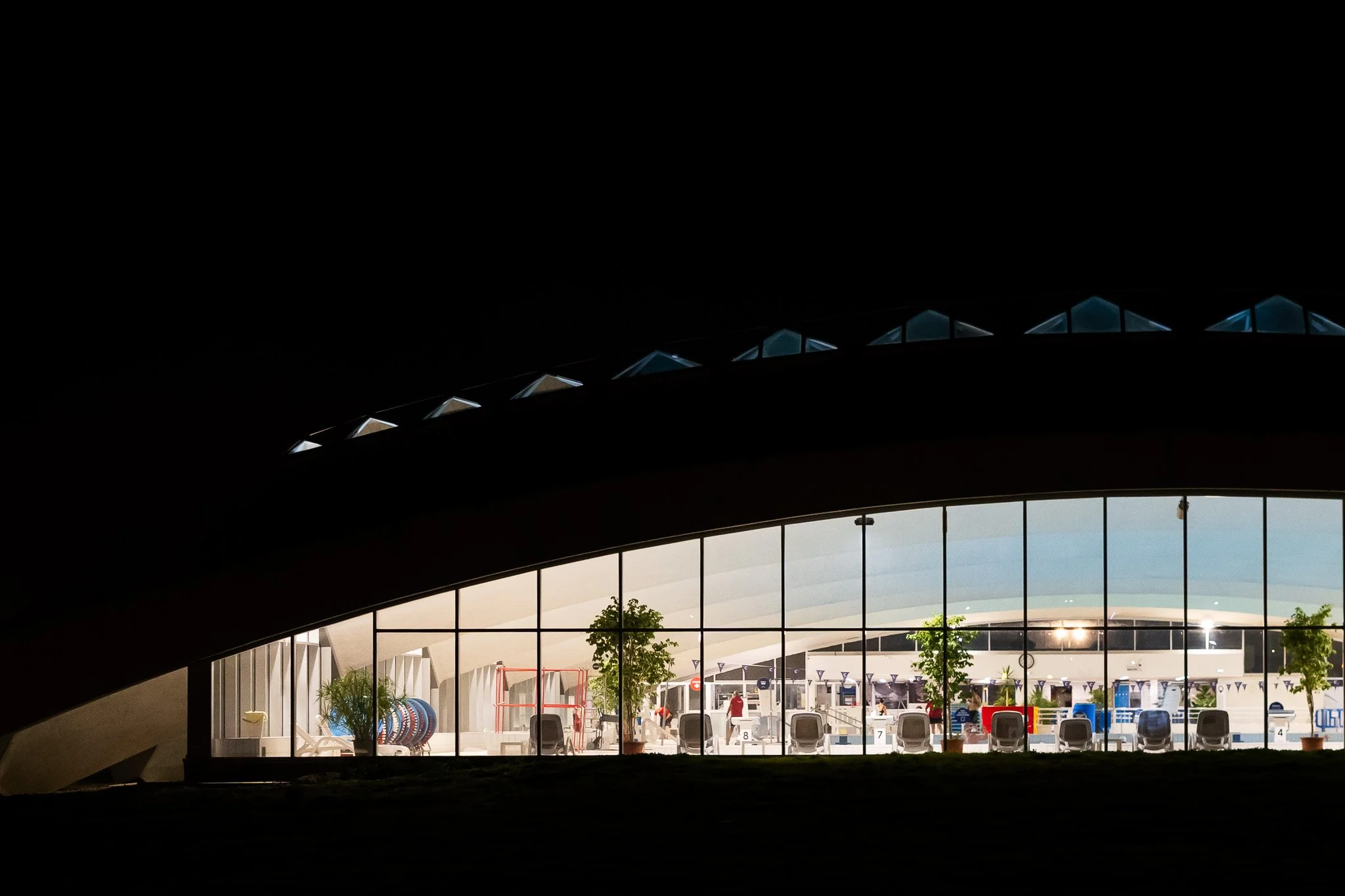 Vue d'un centre aquatique moderne avec grande baie vitrée montrant une piscine extérieure, des chaises et des arbres, prise depuis un espace intérieur sombre.