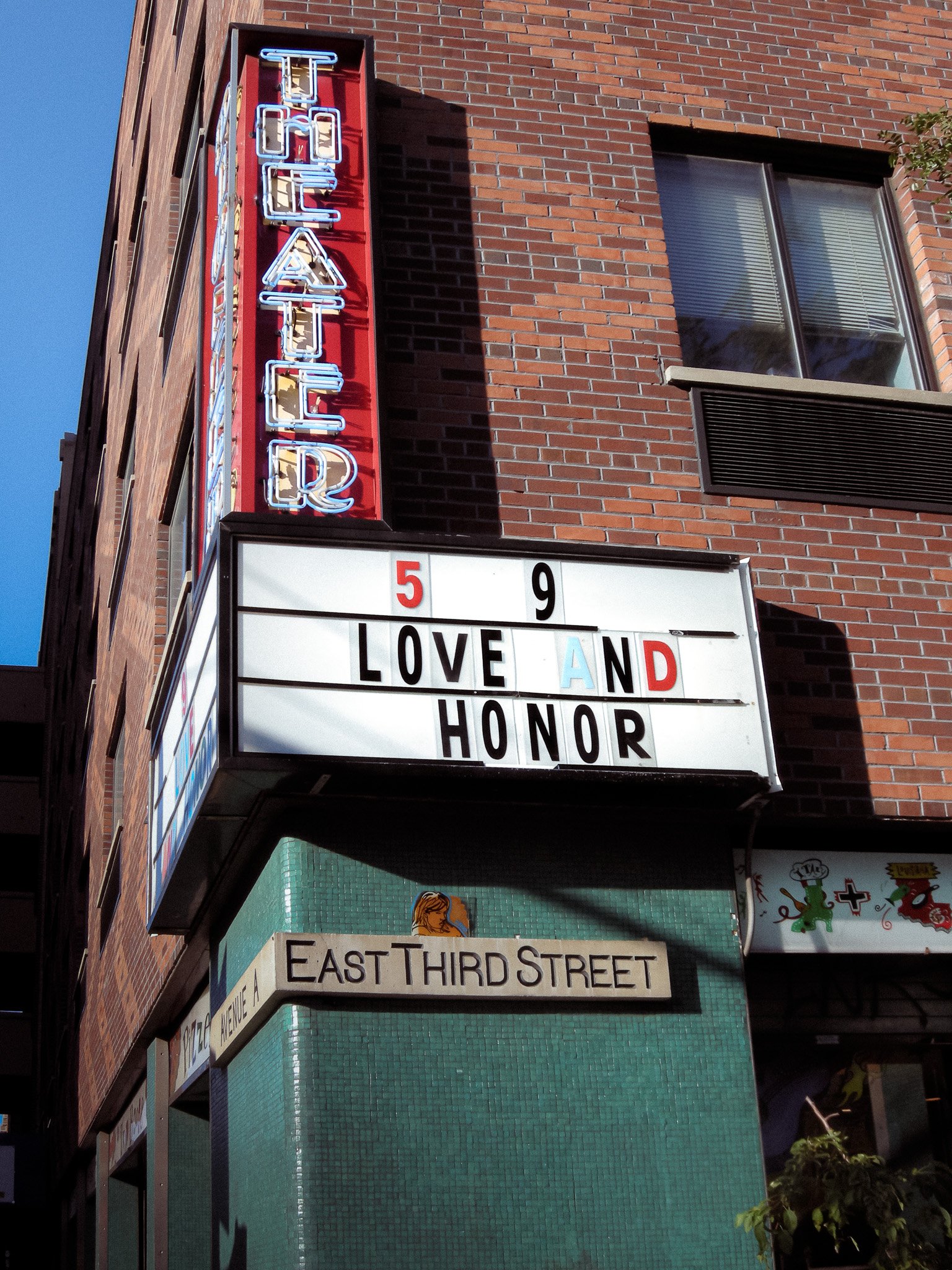 Un panneau lumineux indique le nom d'un établissement, en dessous un enseigne mentionne 'LOVE AND HONOR'. La façade est en briques avec une fenêtre, et une autre enseigne indique 'EAST THIRD STREET'.