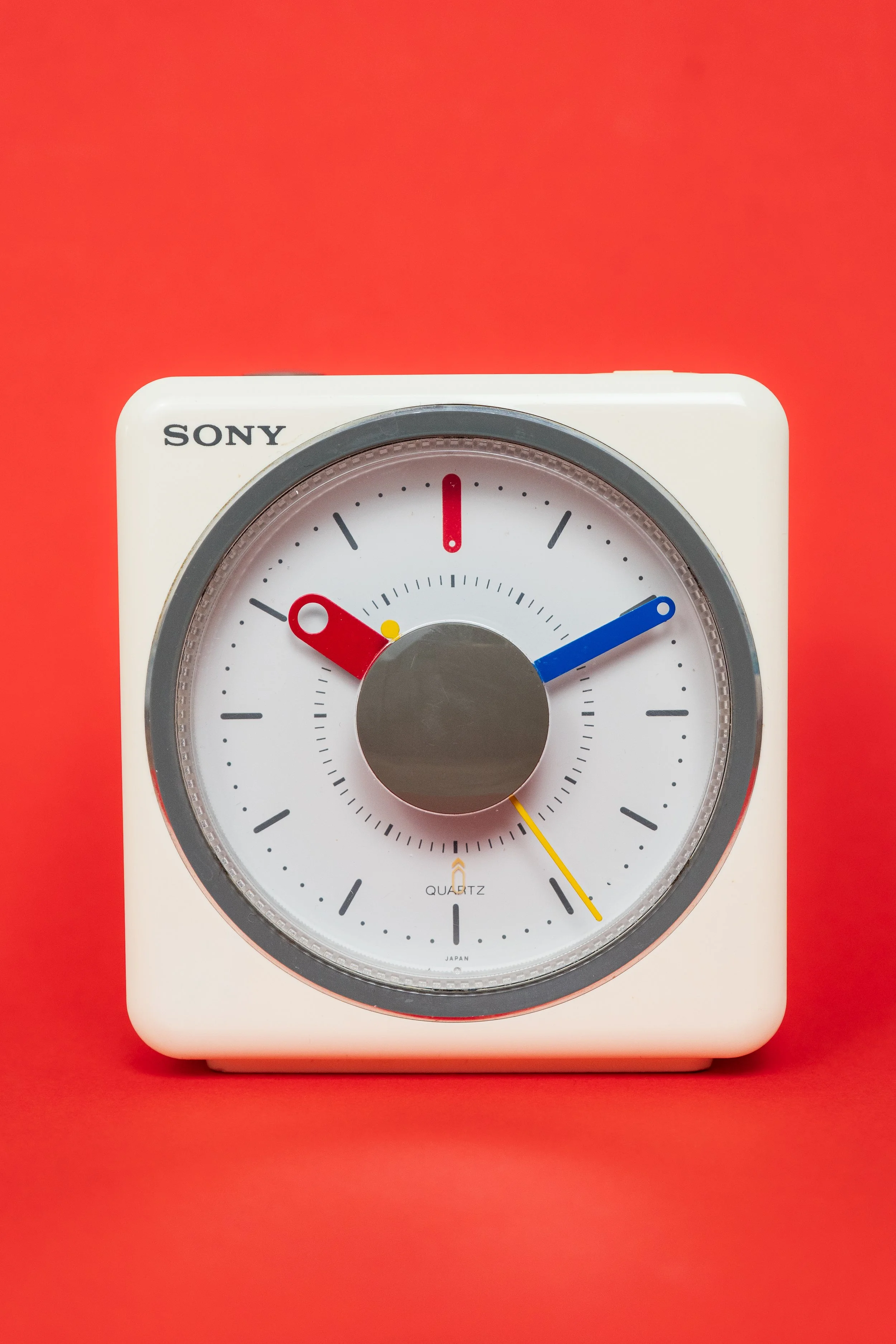 Une horloge analogique blanche avec un cadran noir, une aiguille bleue indiquant 2 heures, une aiguile rouge indiquant 11 heures et une aiguille jaune indiquant environ 4 heures, posée sur un fond rouge.