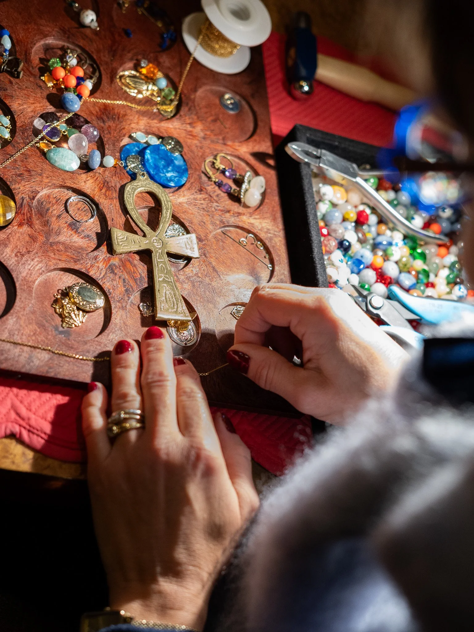 Une personne utilisant une pince pour manipuler des bijoux sur une table en bois, avec divers accessoires et fioles de perles de différentes couleurs entourant la scène.
