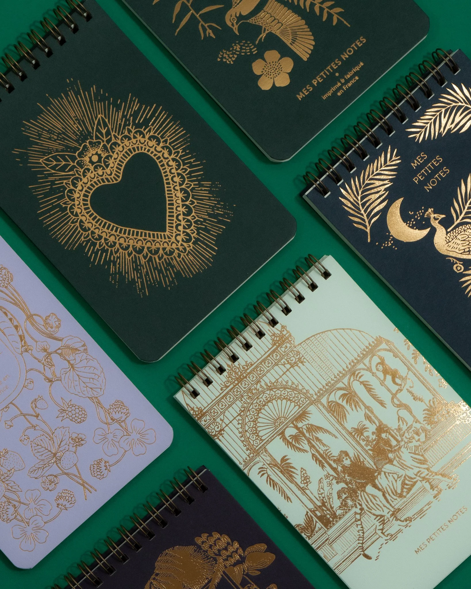 Des carnets de notes avec des couvertures élégantes en couleurs sombres et des motifs dorés, dont un motif de cœur avec des rayons lumineux, un motif floral, un motif de pagode japonaise, et un motif de signe celtique, tous liés à un style artistique ornemental.