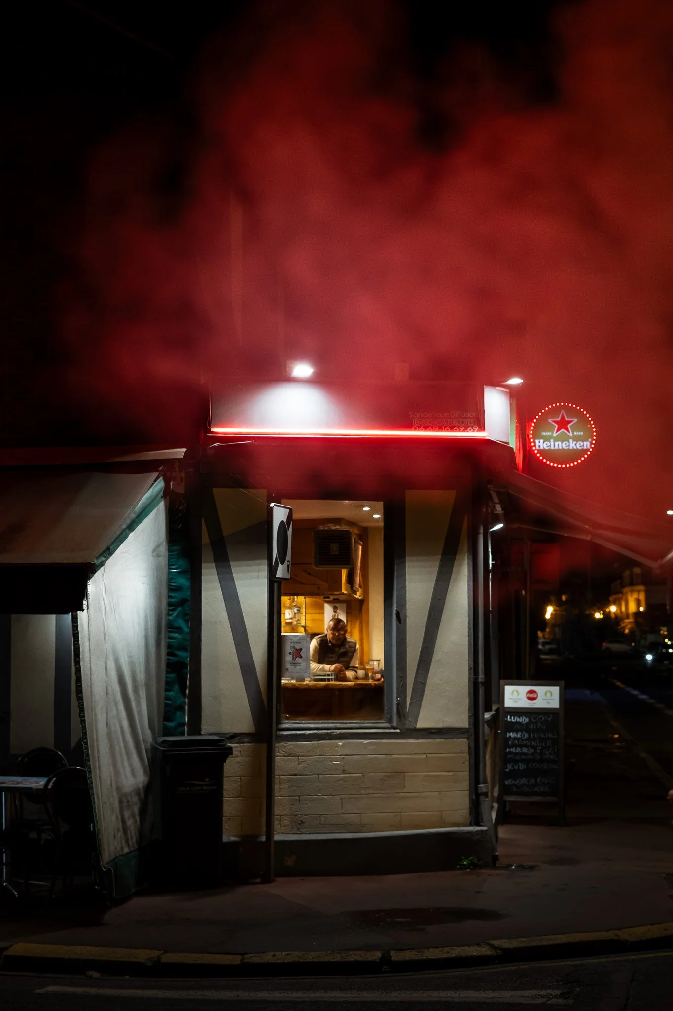 Bar avec une personne à l'intérieur, enseigne lumineuse Heineken et menu écrit à la craie à l'extérieur, scène nocturne.