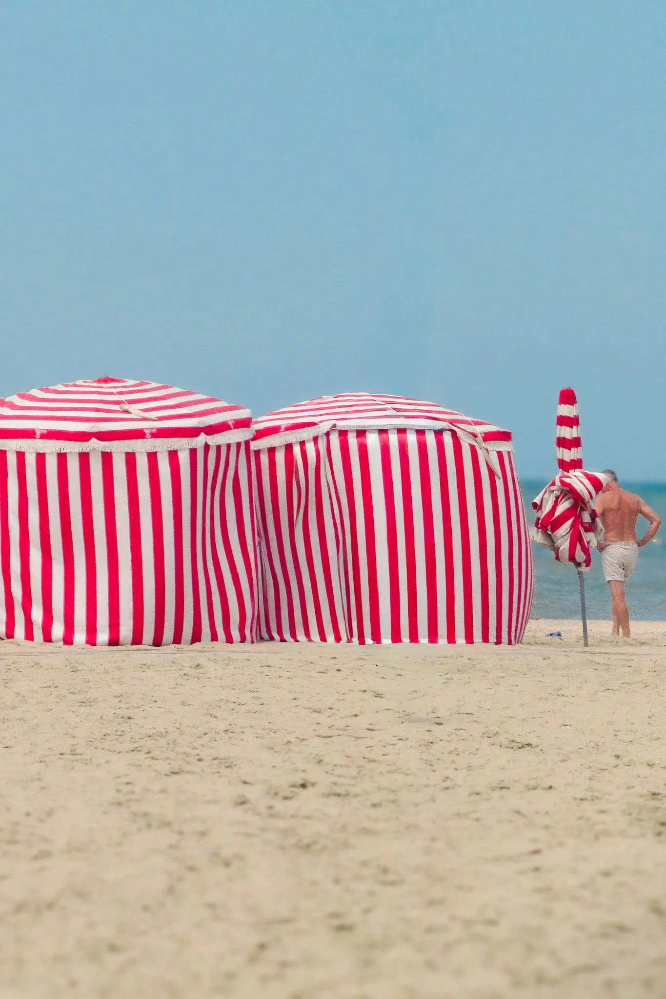 Plage avec des parasols à rayures rouges et blanches, avec un ciel bleu clair, plage de sable clair, et une personne à droite à côté d'un parasol.