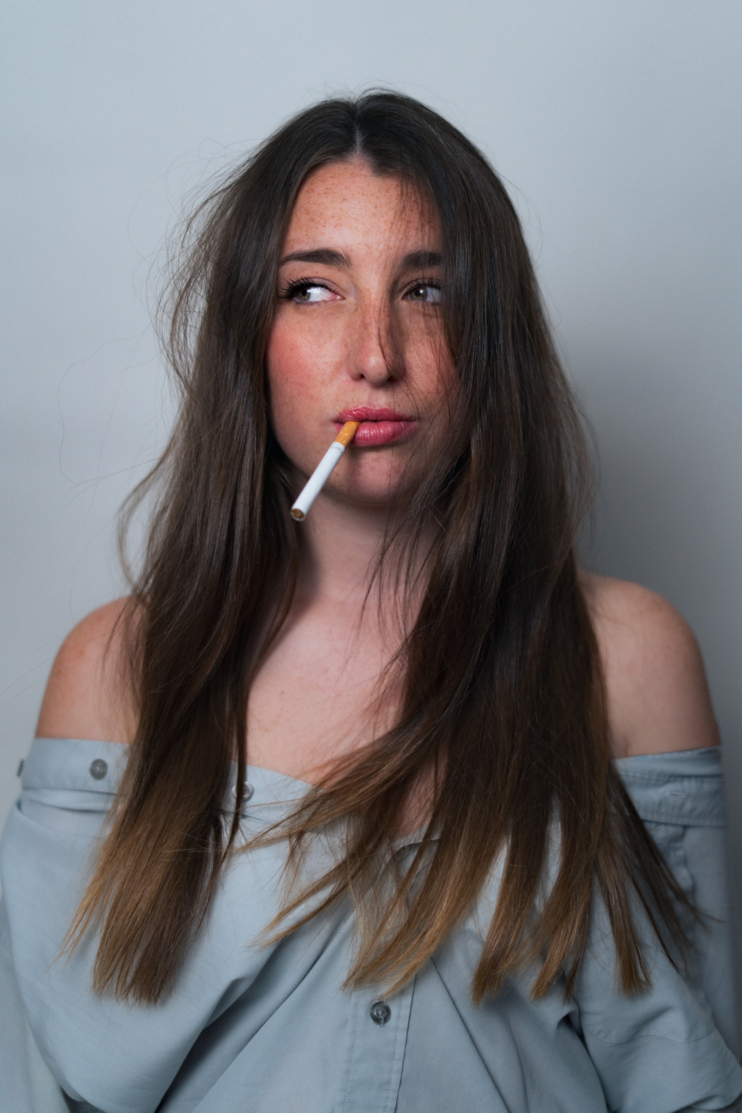 Jeune femme avec de longs cheveux châtains, des taches de rousseur, portant une chemise grise, avec une cigarette entre les lèvres, regarde sur le côté, fond gris clair.