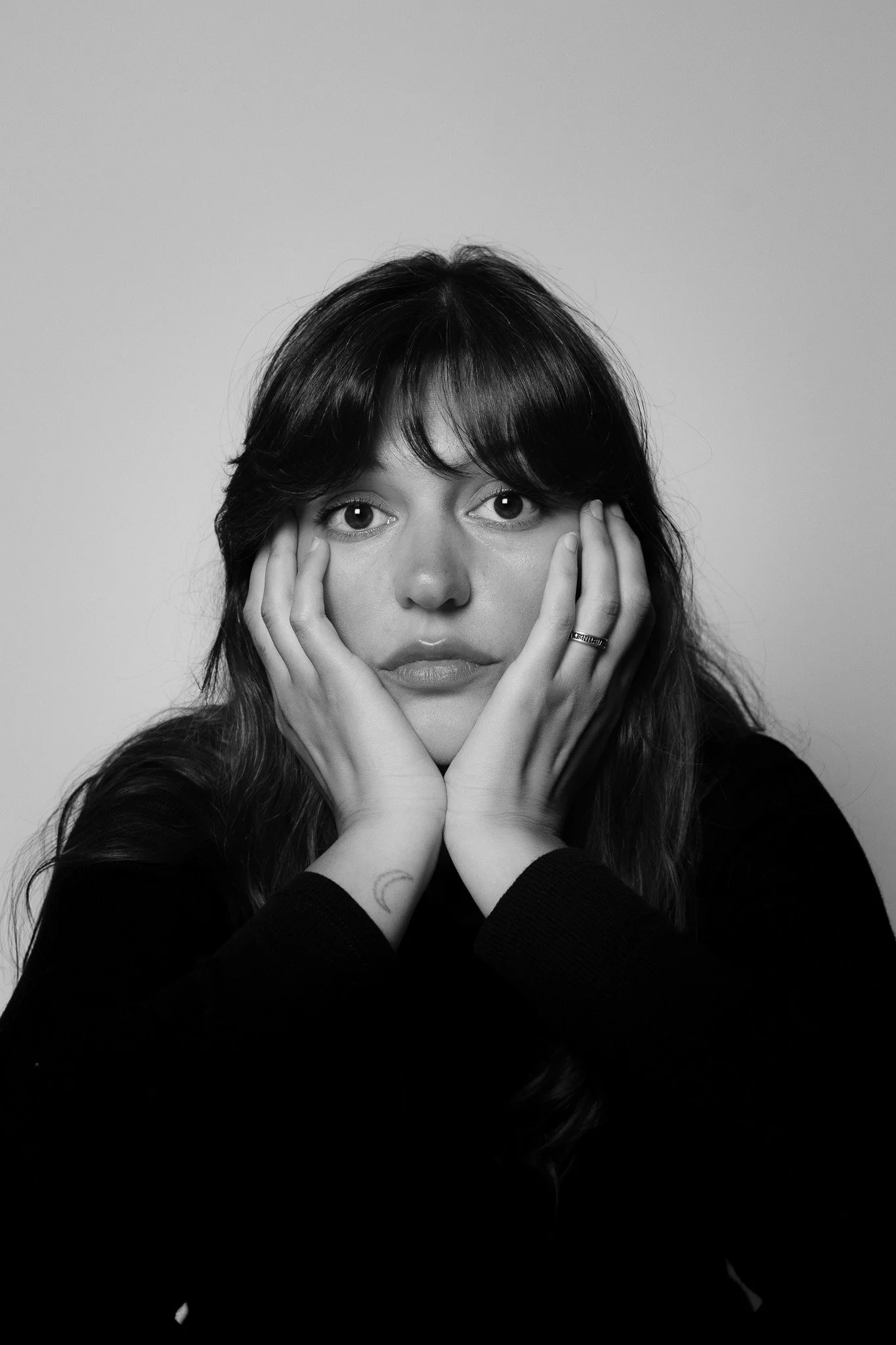 Une femme avec des cheveux bruns, portant un pull noir, pose avec les mains sur son visage, regardant devant elle. La photo est en noir et blanc.
