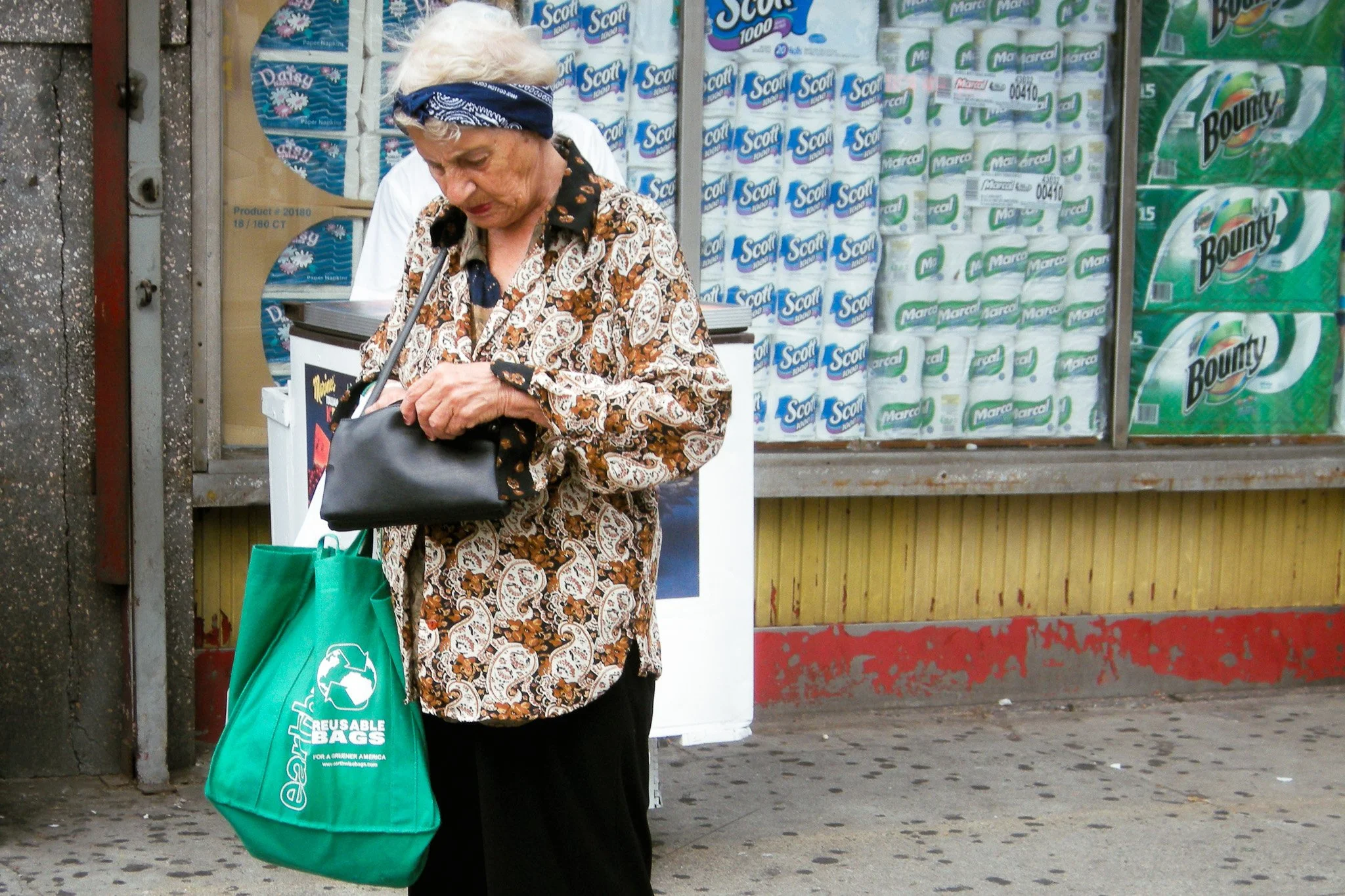 Une femme âgée avec des cheveux gris portant un bandana bleu, un chemisier à motifs, portant un sac en toile de recharges d'énergie, se tient devant un étalage de produits de papier toilette dans une zone urbaine.