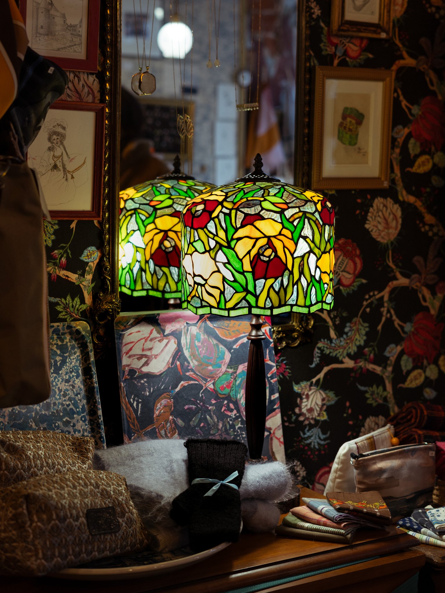 Intérieur tamisé d'une boutique avec une lampe Tiffany coloré avec un motif floral, entouré de cadres avec des illustrations et des décorations textiles vintage.