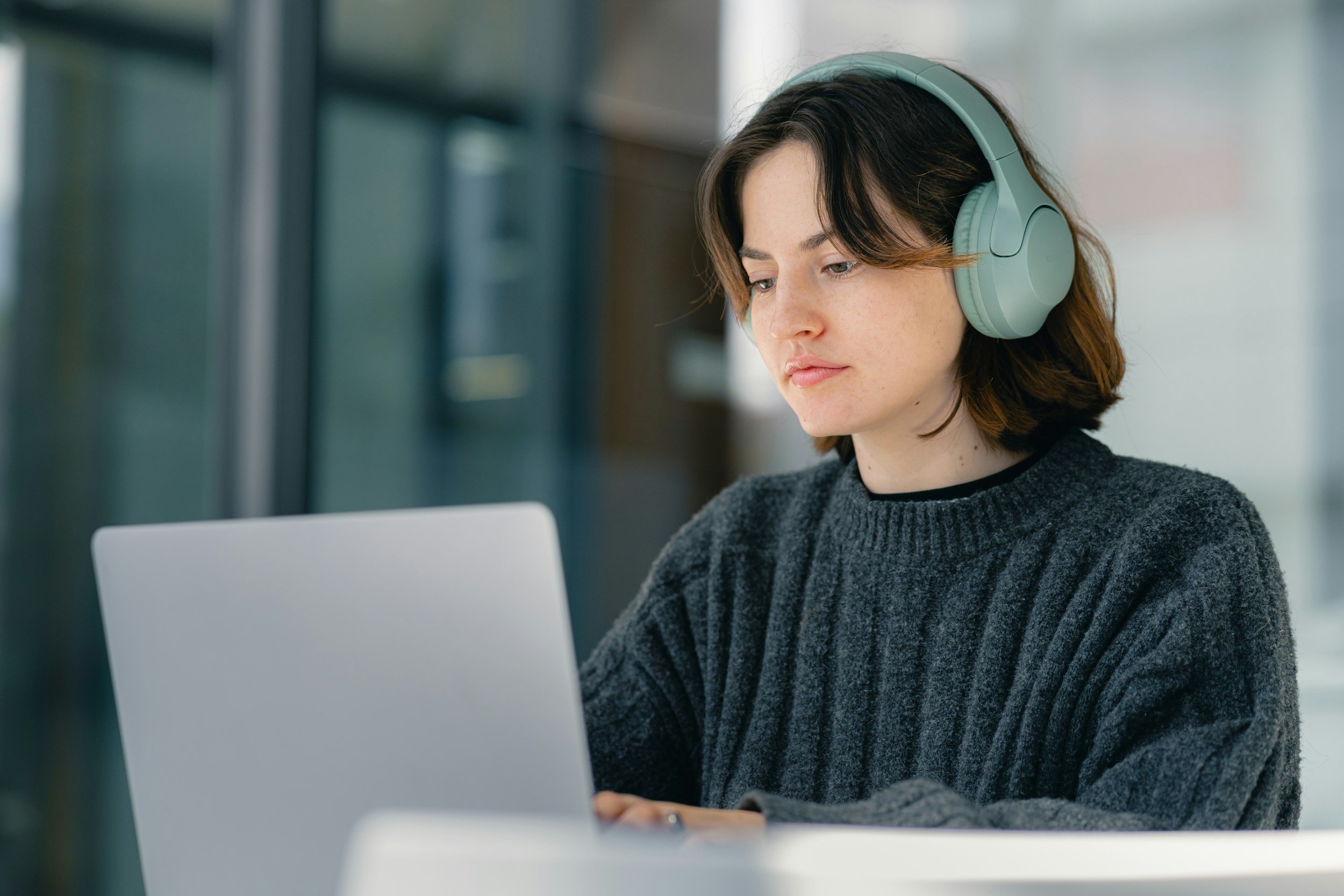 A young woman with short brown hair, wearing a dark gray sweater and mint green over-ear headphones, appears focused while working on a silver laptop in a modern bright indoor setting.