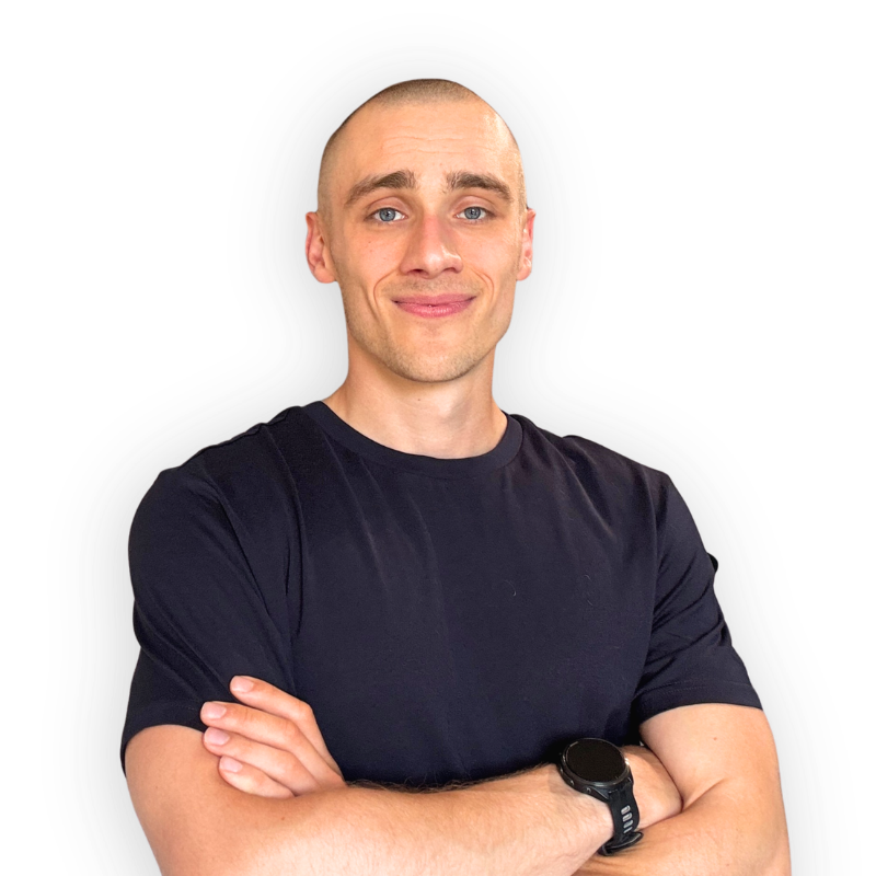 A young man with a shaved head and blue eyes, wearing a black T-shirt and a watch on his left wrist, smiling with arms crossed against a plain background.