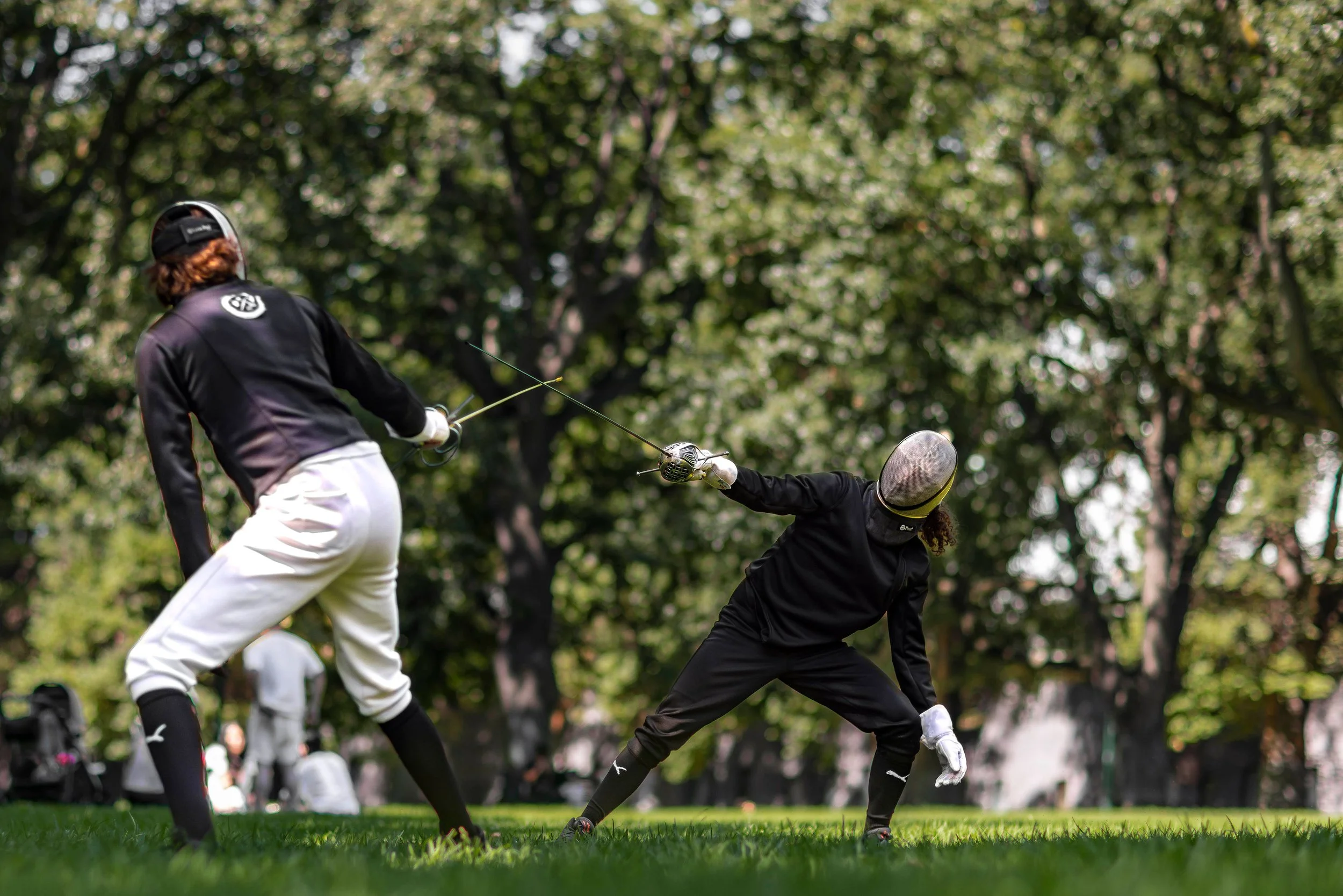 Coaches Woody & Habibi are fencing in the Carlton Gardens park, wearing full fencing gear including masks and jackets. Trees and grass are visible in the background.