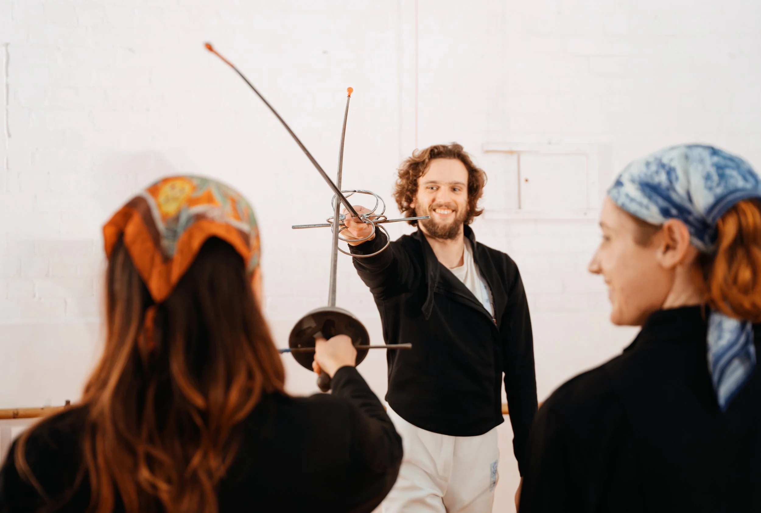 Woody demonstrating some simple rapier fencing techniques to two engaged students wearing headscarves, interested in HEMA.