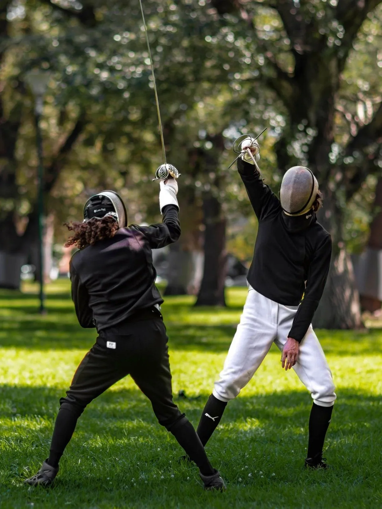 Coaches Woody & Habibi are fencing in the Carlton Gardens park, wearing full fencing gear including masks and jackets. Trees and grass are visible in the background.