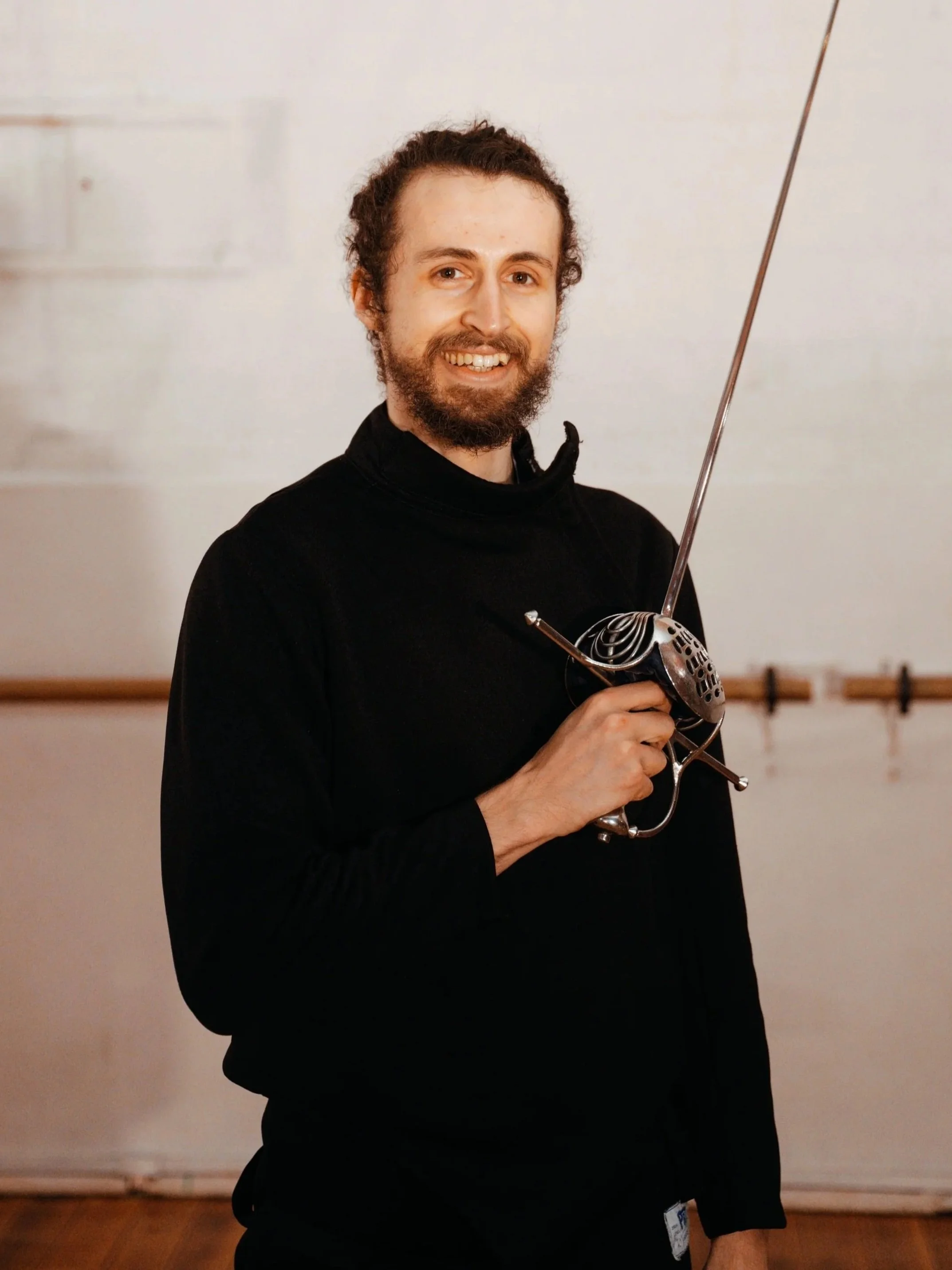 Head Instructor Habibi Favilla of Melbourne Historical Fencing is smiling with his Rapier against a white wall.